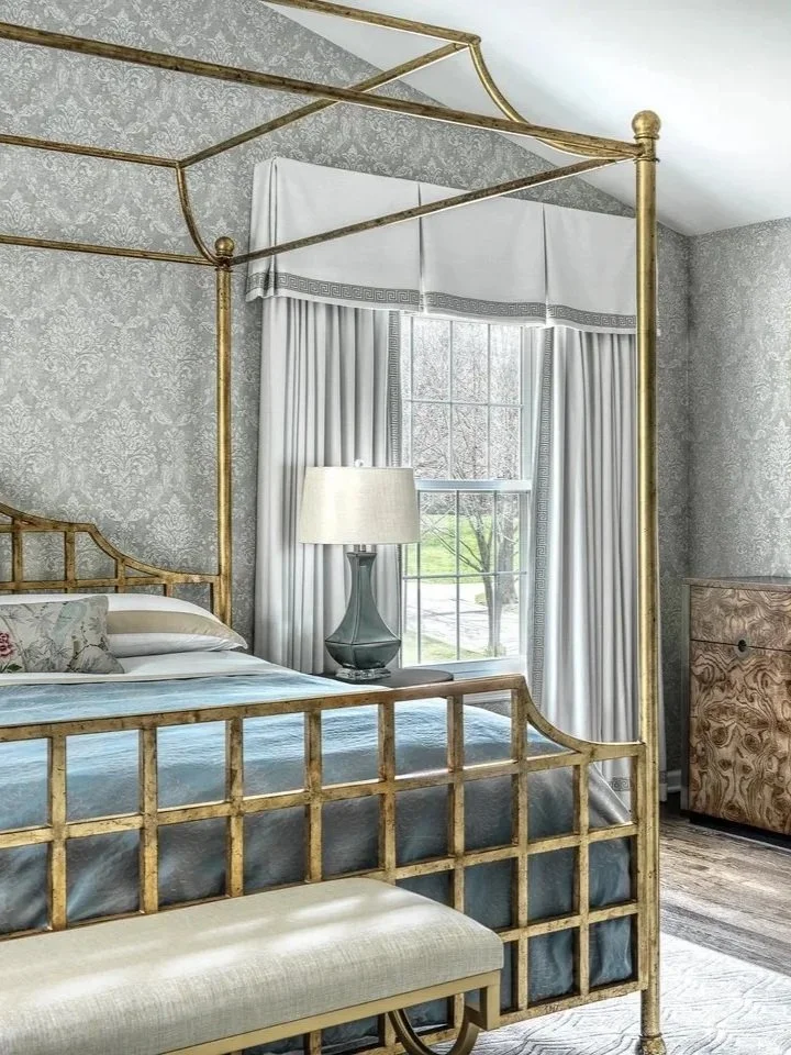 Elegant bedroom with a gold canopy bed, white curtains, a window with a view of a tree outside, a lamp on a nightstand, and a wooden dresser.