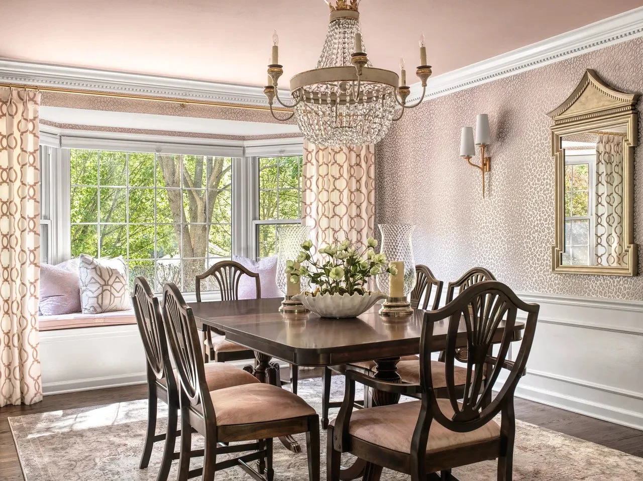 Elegant dining room with a dark wooden dining table surrounded by matching chairs with upholstered seats, a large window with patterned curtains, a crystal chandelier hanging above, a decorative mirror on the wall, and a window seat with cushions by the window, with greenery outside.