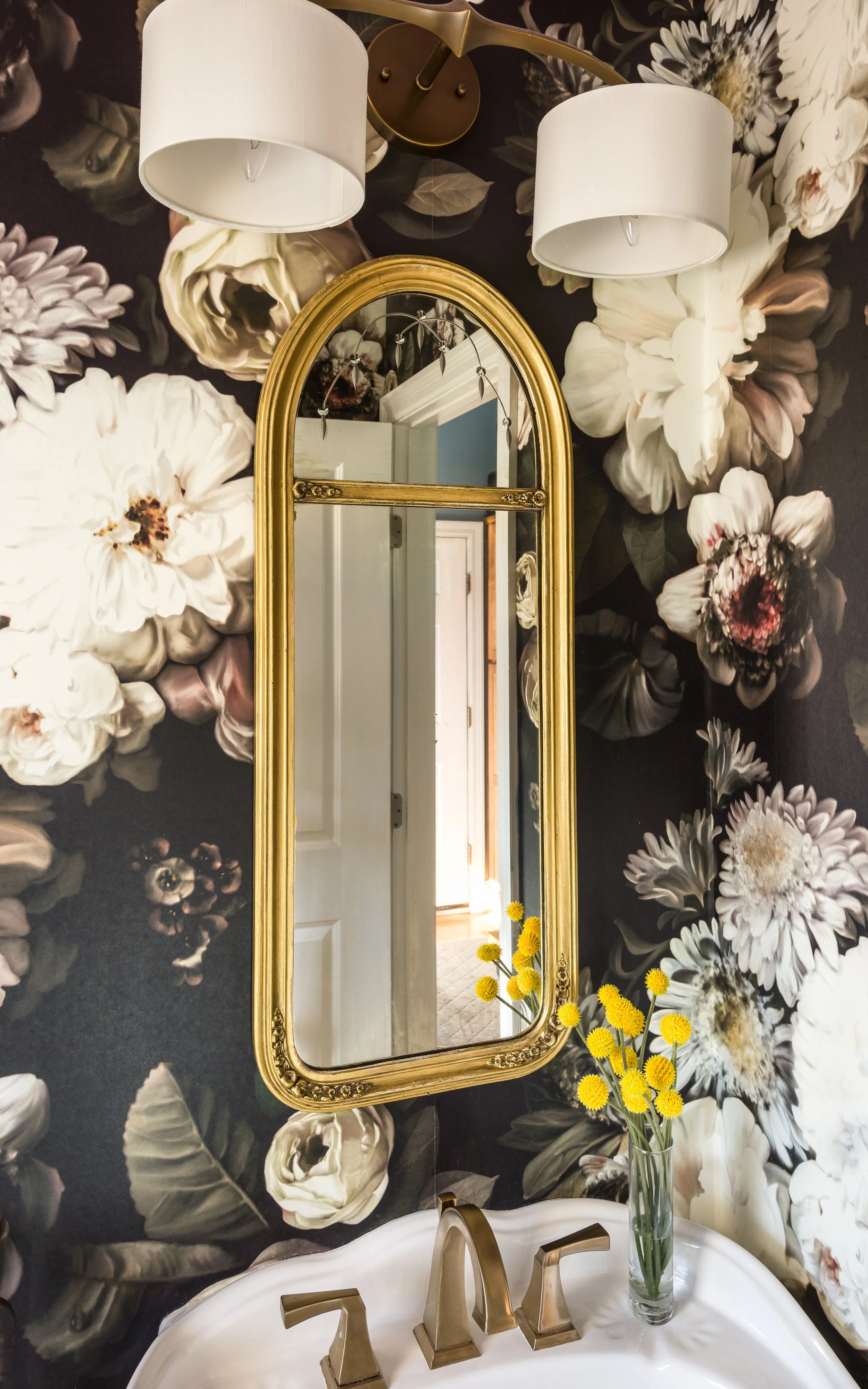A bathroom mirror with a gold frame and a vase with yellow flowers on a white sink. The wall behind features large black, white, and beige floral wallpaper. Two white lamps are mounted above the mirror.