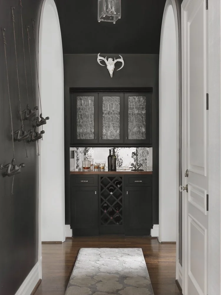 A hallway leading to a black wet bar area with glass-front cabinets, a wine rack, and a sink. A deer skull with antlers is mounted on the dark wall above the bar. The floor is wood with a light-colored rug.