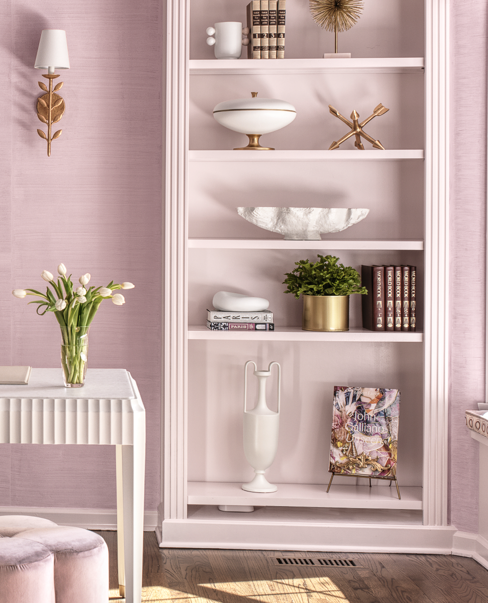 Pink wall with a built-in bookshelf filled with decorative items, books, and plants, next to a white desk with a vase of tulips and a pink cushioned stool.