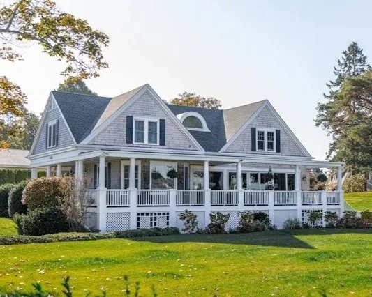 A large white house with a wraparound porch, multiple gabled roofs, and dark shingles, surrounded by green trees and a well-maintained lawn.