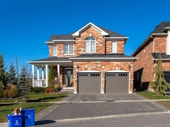 A two-story brick house with attached two-car garage on a clear sunny day, with a paved driveway, small front lawn, and neighboring house to the right.