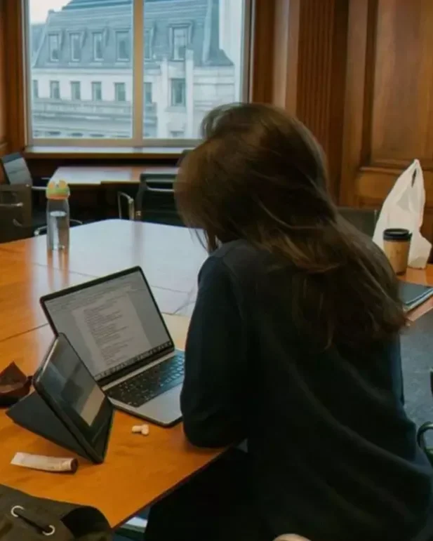 A woman with brown hair is sitting at a wooden table in a room with wood-paneled walls, working on a laptop. There is a tablet, a coffee cup, and some small items on the table. A window behind her shows a building outside.