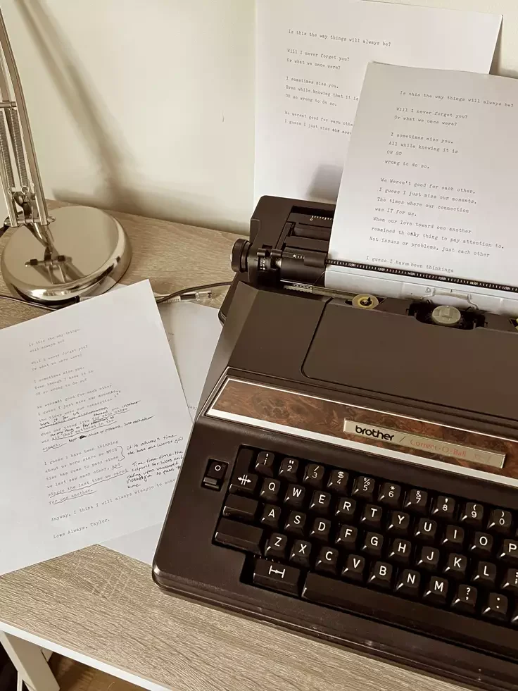A vintage black Brother typewriter with typed pages and handwritten notes on a wooden desk next to a modern silver desk lamp.