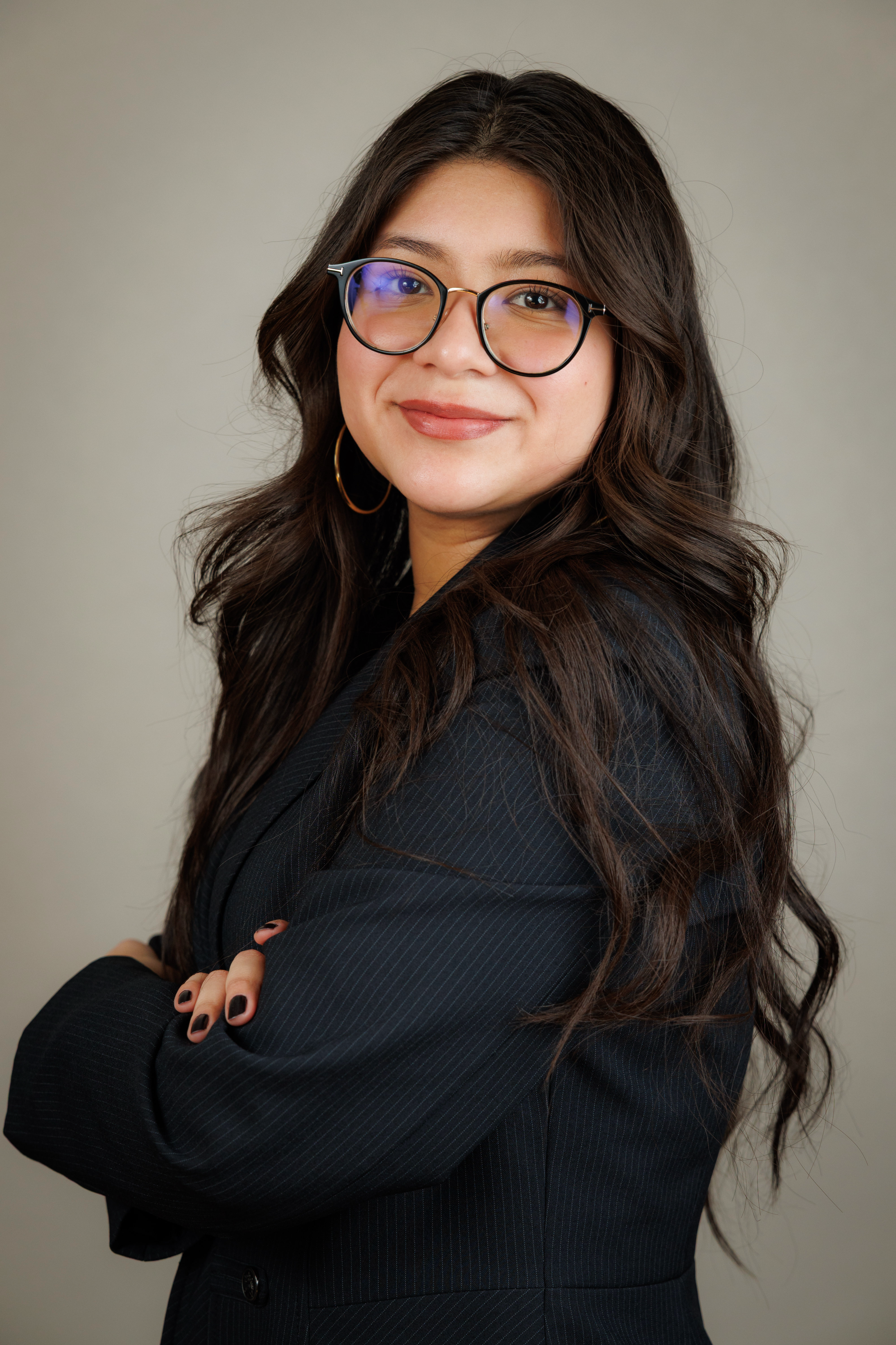 A young woman with long, wavy dark hair wearing large glasses with blue light reflection, hoop earrings, and a black pinstripe blazer, standing against a neutral background with her arms crossed and smiling at the camera.