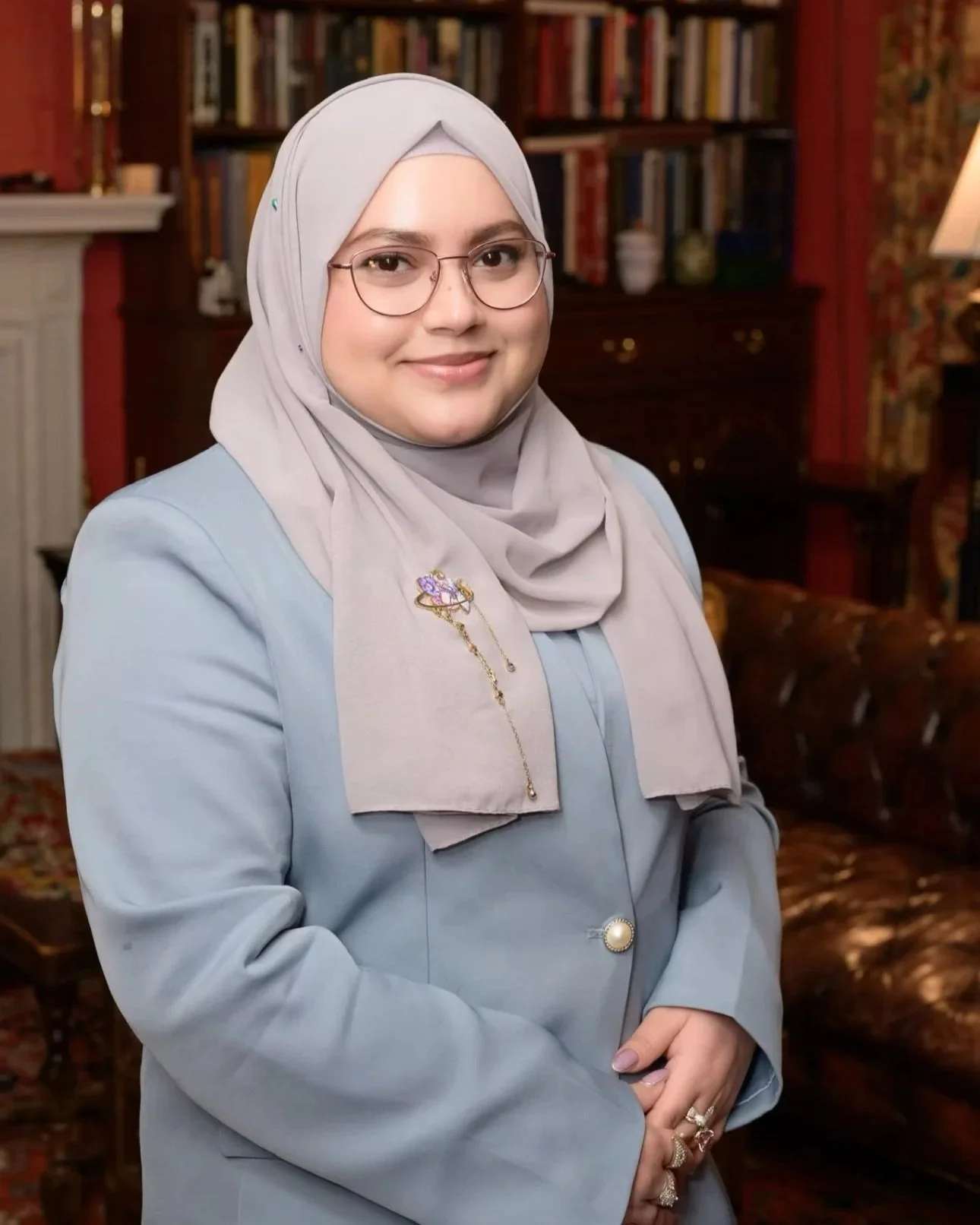 A woman wearing a light gray hijab and glasses, dressed in a light blue suit, standing in a room with wooden bookshelves and a fireplace in the background.