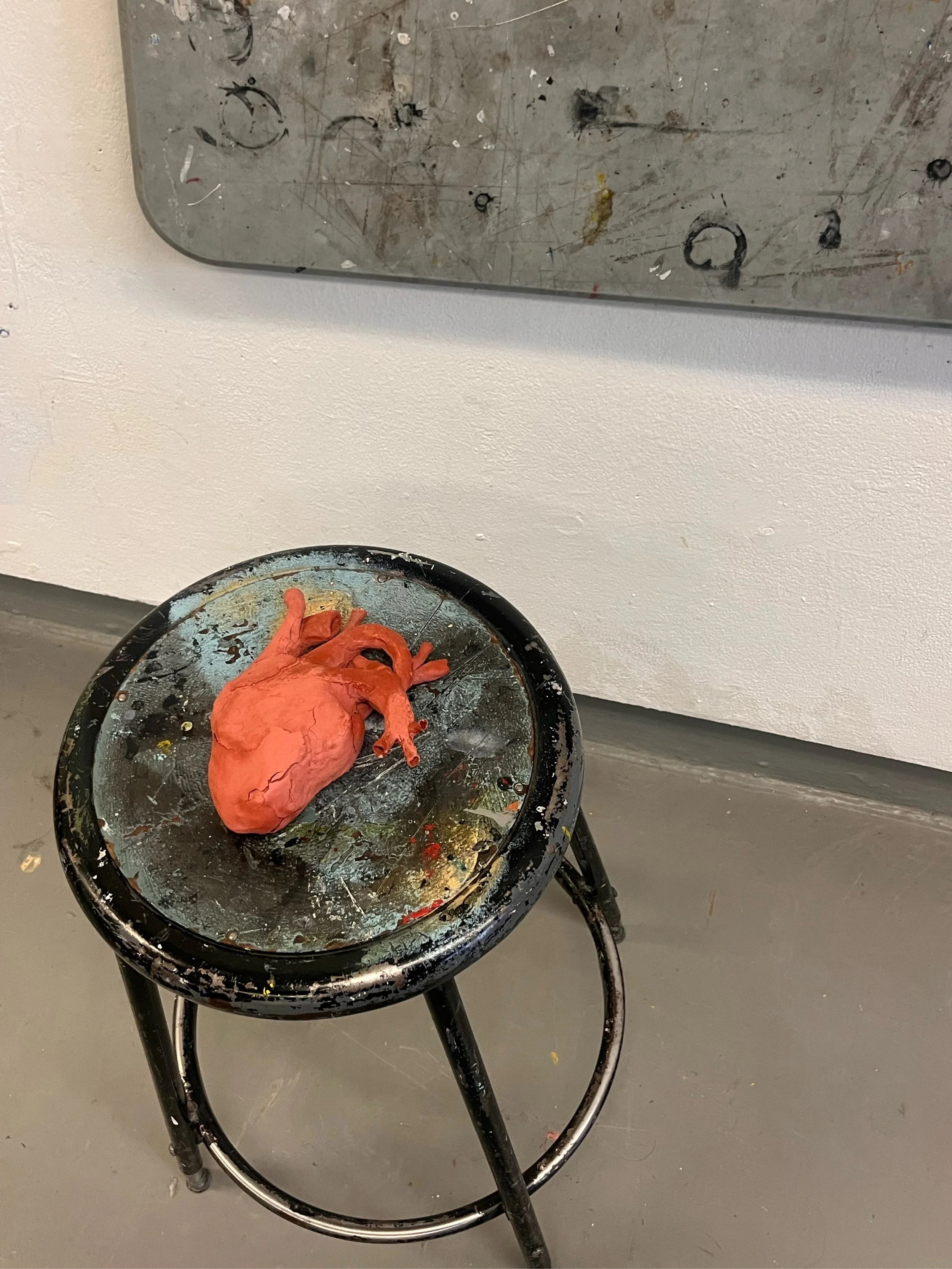 A small black metal stool with a worn paint surface, holding a red anatomical model of a human heart. In the background, there is a gray wall and a metallic surface with paint splatters and scribbles.