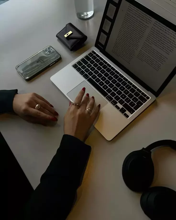 Person working on a laptop at a desk with a water bottle, cell phone, wallet, headphones, and glasses.
