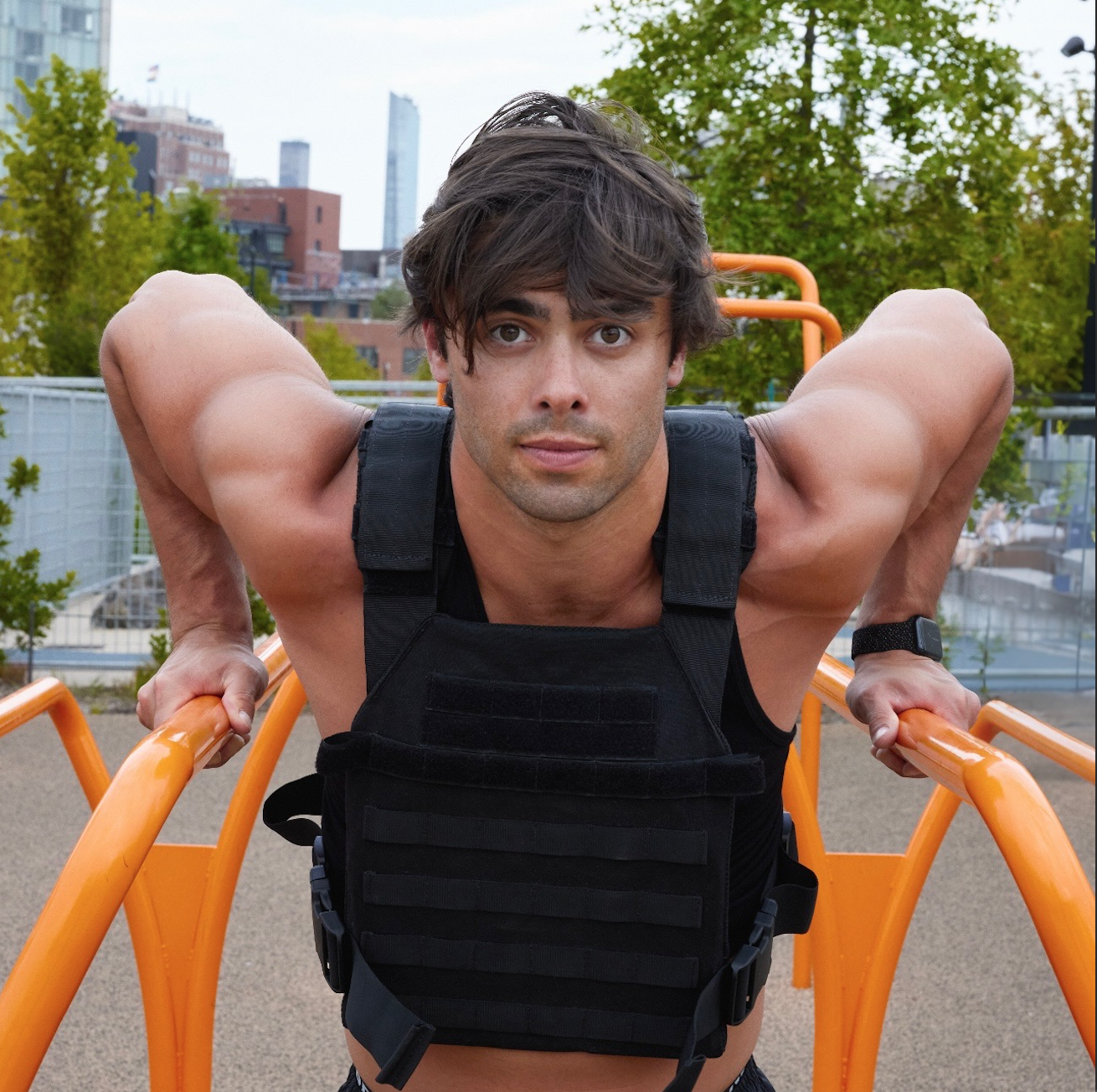 A young man doing push-ups on orange outdoor exercise equipment in a park, with trees and city buildings in the background.