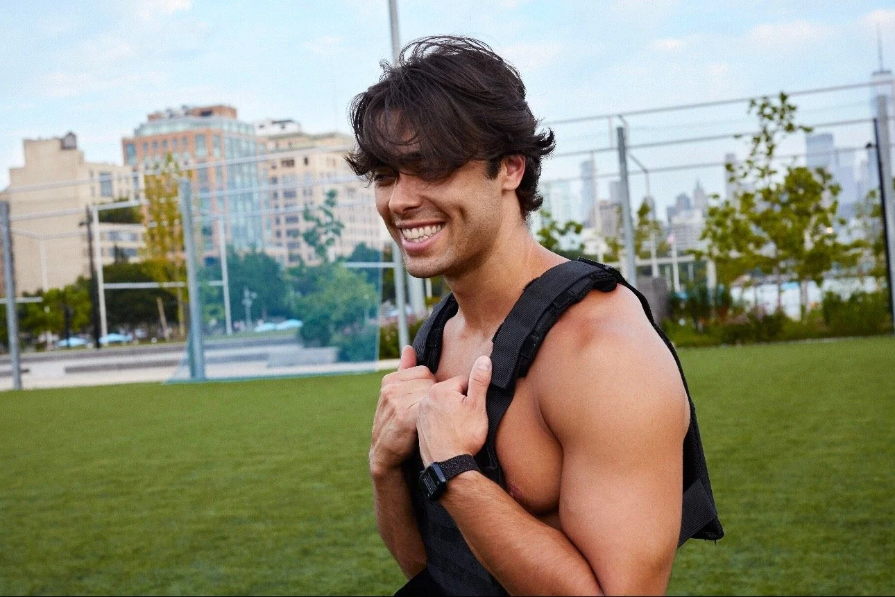 A smiling young man with dark hair, shirtless, outdoors on a grassy field, wearing a black backpack and a watch, with city buildings in the background.