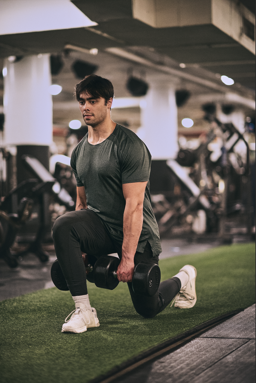 A man in a black athletic shirt and pants performing a lunge exercise with a dumbbell in a gym.