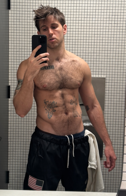 A shirtless young man with tattoos taking a mirror selfie in a bathroom with tiled walls.