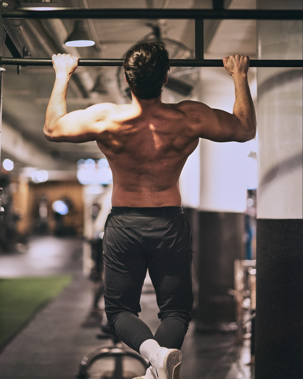 Young man doing pull-ups at a gym, shirtless, wearing black pants and white shoes.