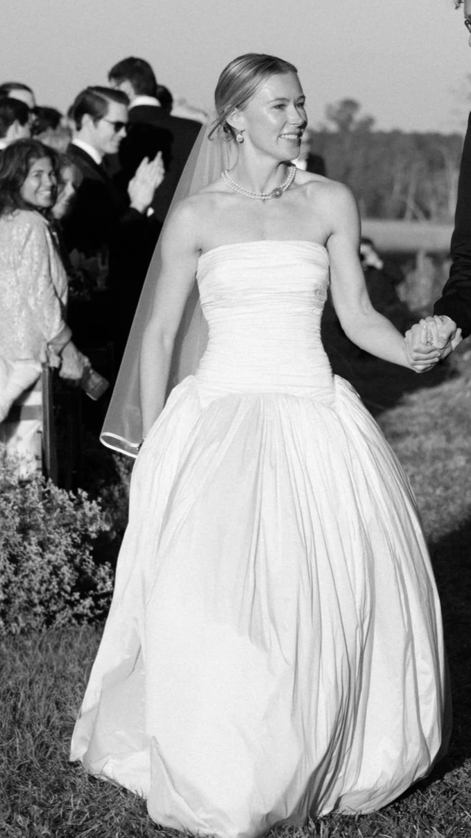 Black and white photo of a smiling woman in a strapless wedding gown holding hands with someone. Several people are visible in the background, some clapping and smiling.