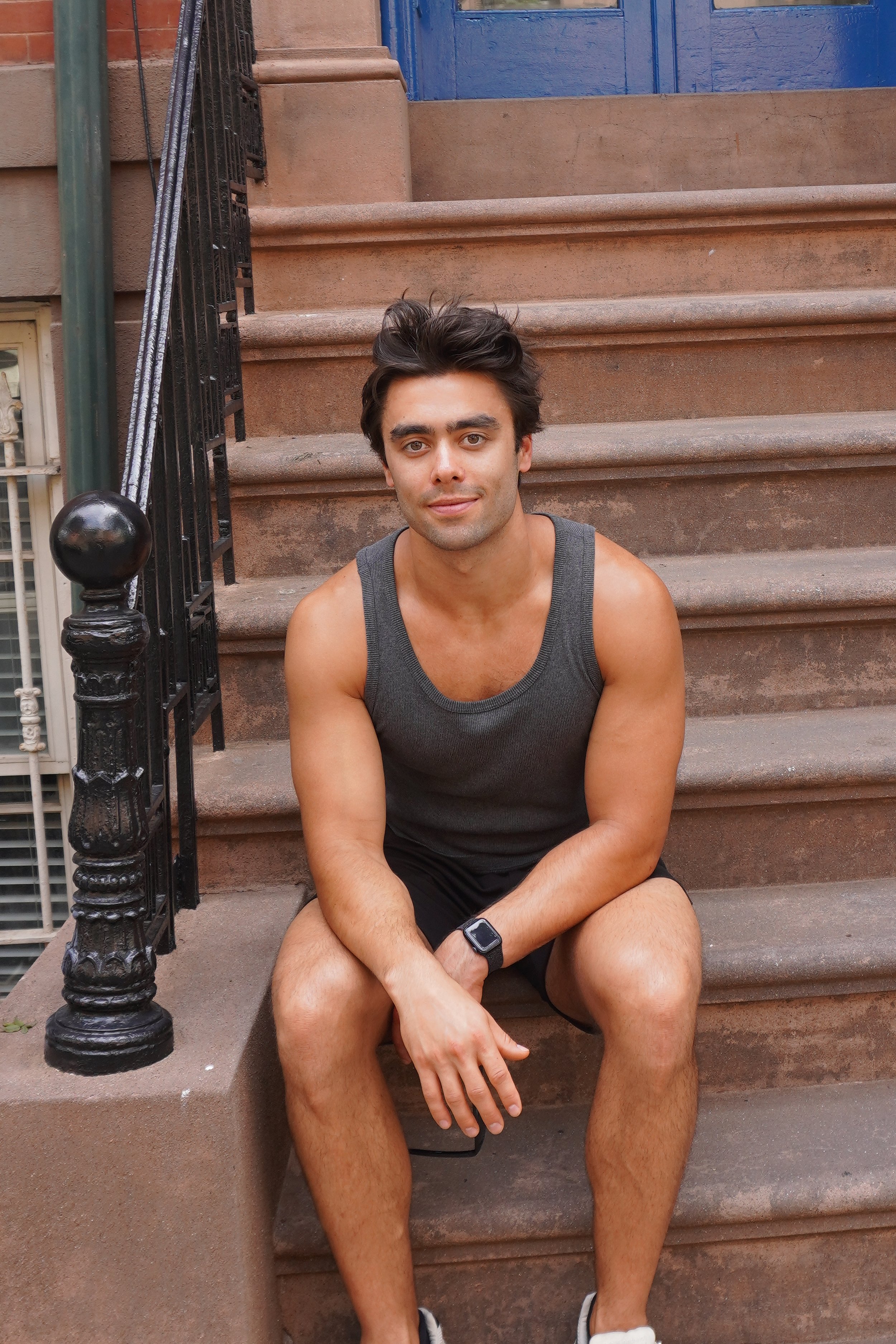 A young man with dark hair, wearing a gray tank top, black shorts, sitting on brownstone stairs with wrought iron railing and a blue door in the background.