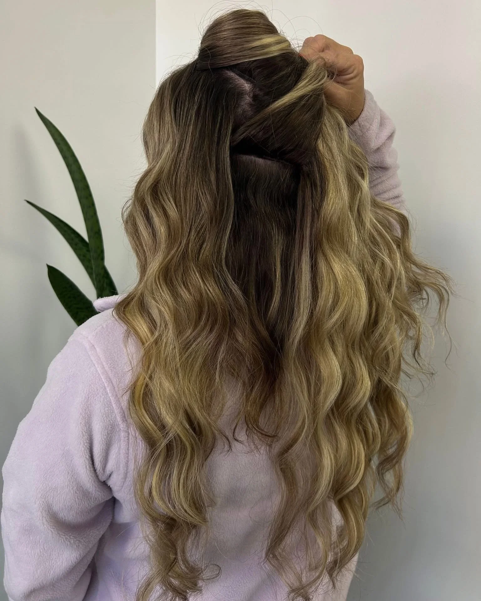 A woman with long, wavy blonde hair wearing a pale pink sweater, standing against a plain light-colored wall.