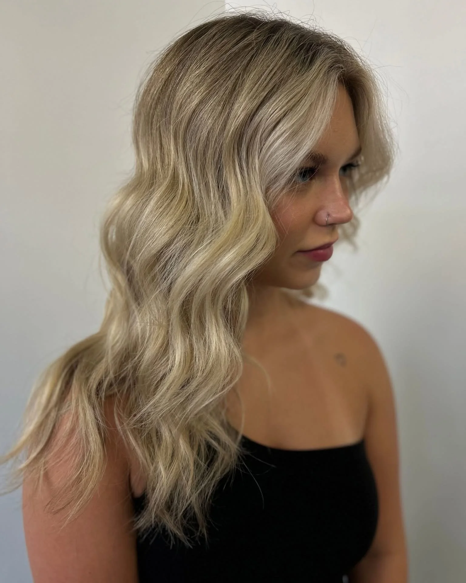A woman with long, wavy blonde hair, a nose ring, and wearing a black strapless top, standing against a plain white wall.