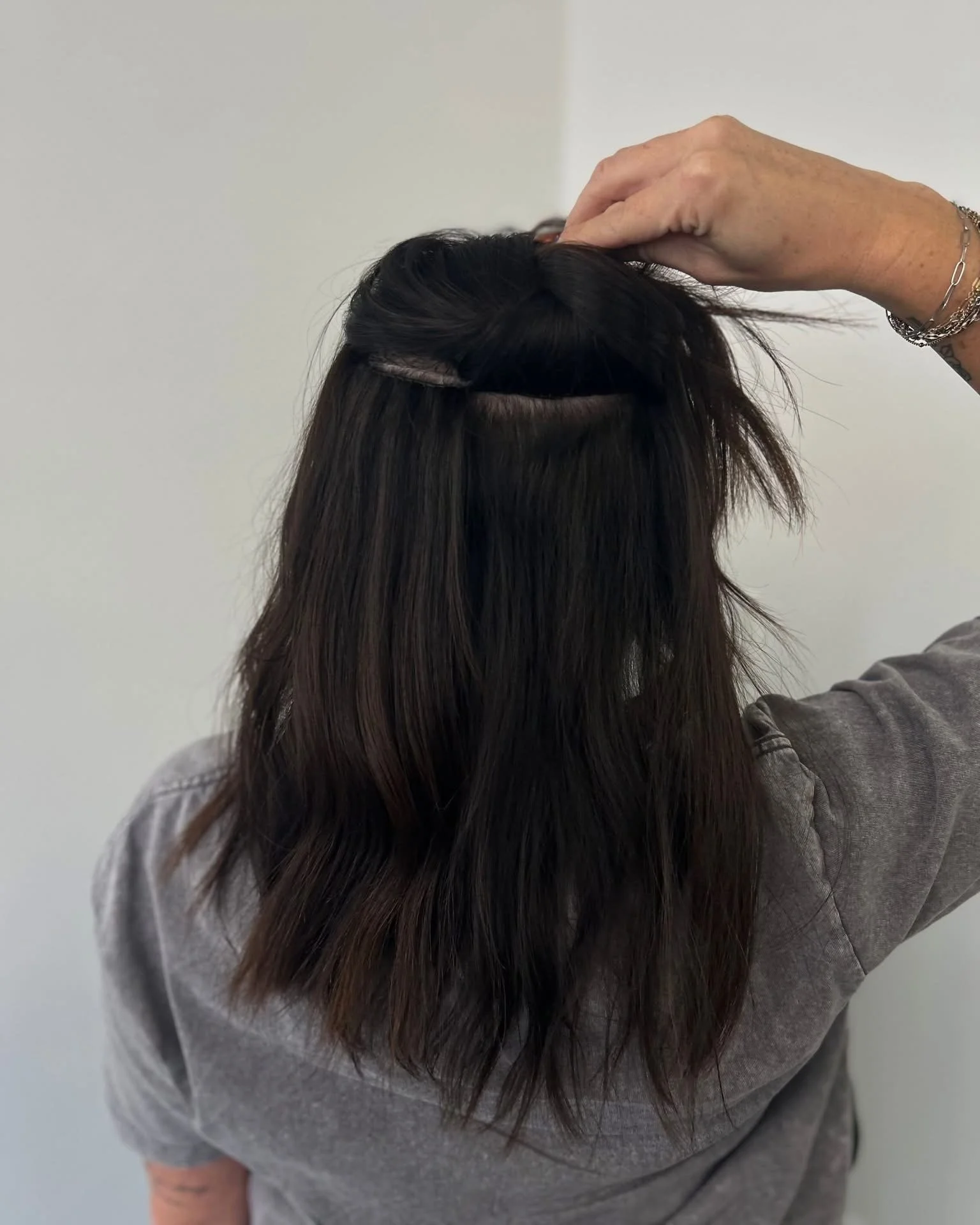 A person with long, dark brown hair wearing a gray shirt, lifting part of their hair to reveal the roots and a hair extension.