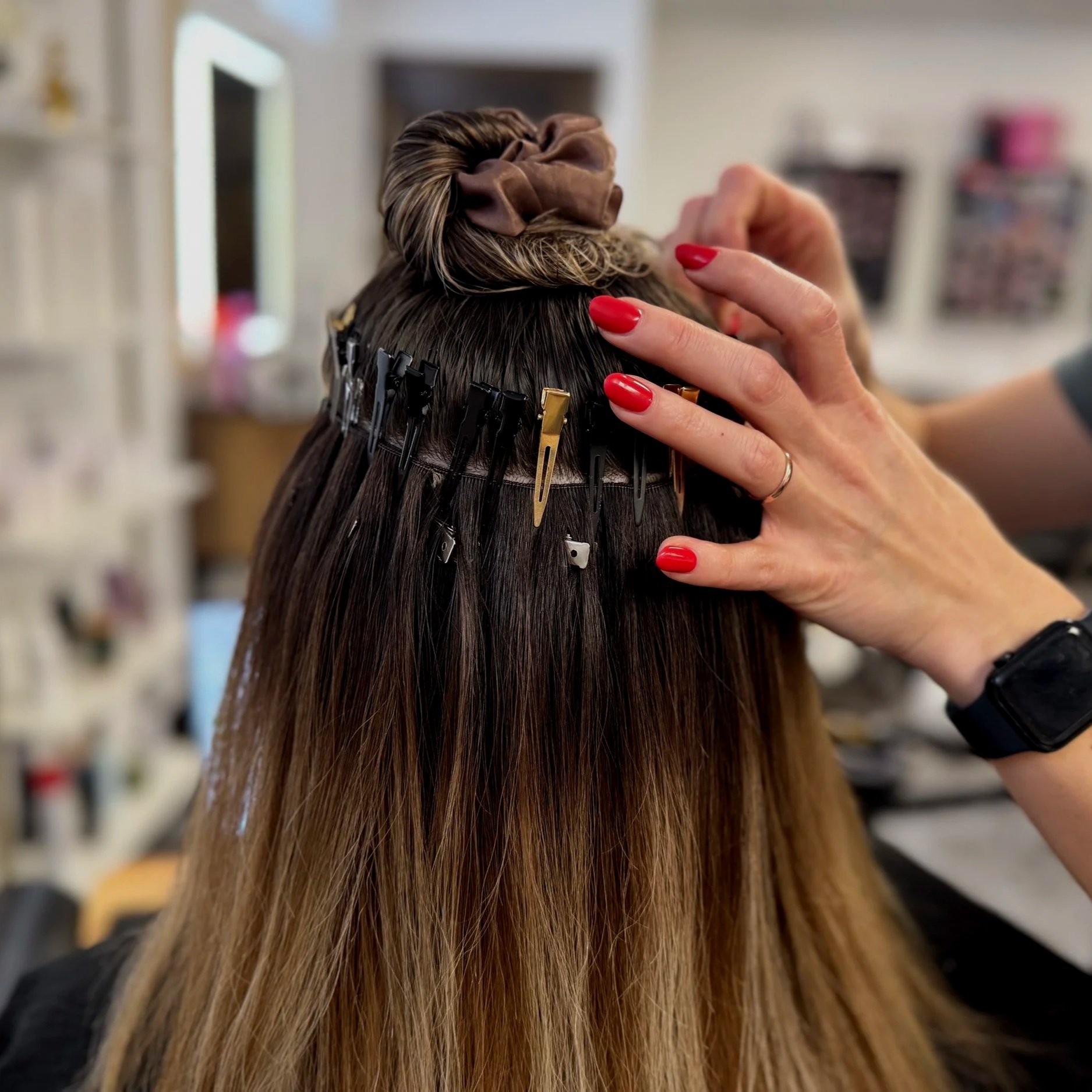 A person with red painted nails is styling brunette hair, clipped in sections with black and gold clips, in a salon setting.