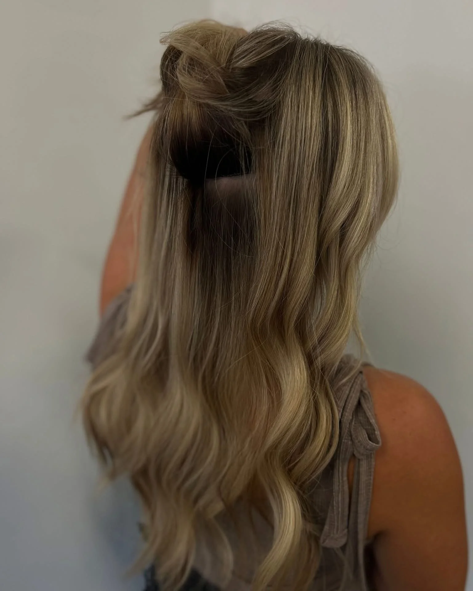 A woman with long, wavy blonde hair wearing a brown sleeveless top, standing against a plain white wall.