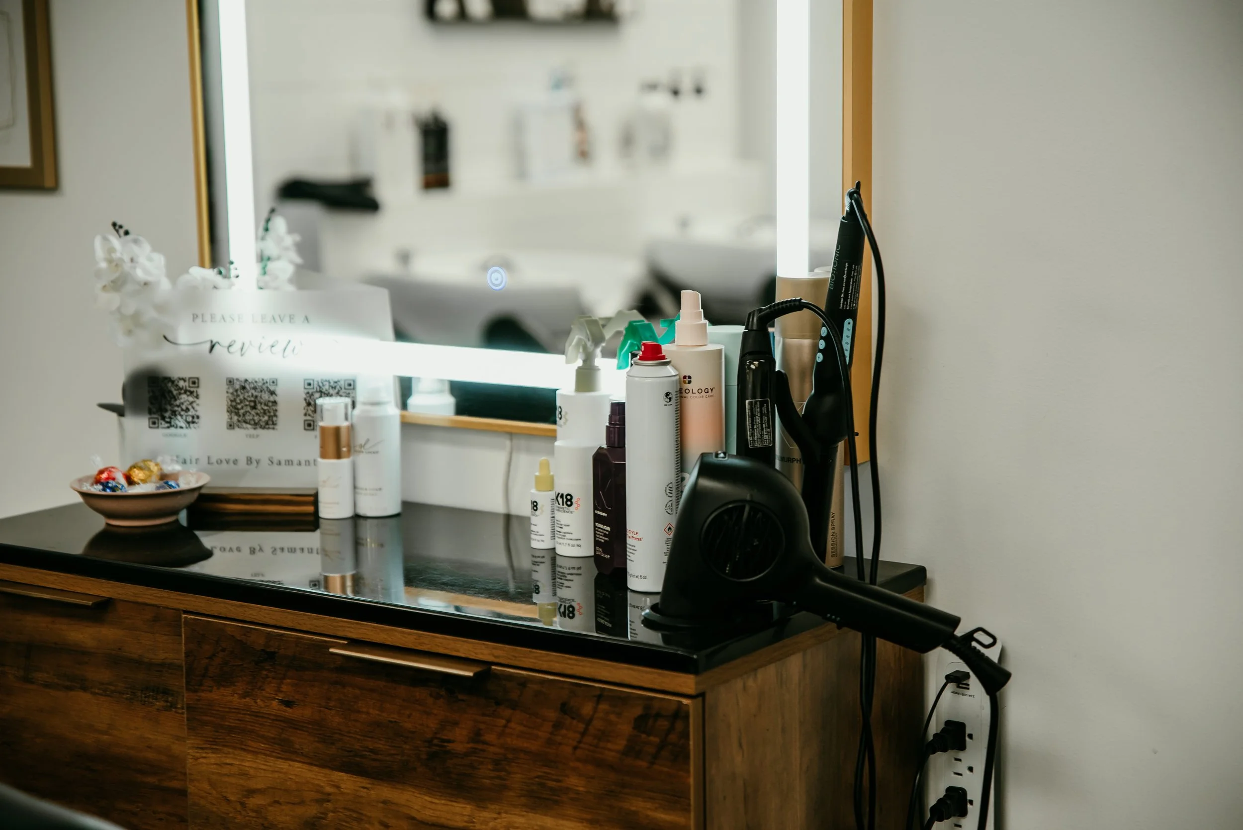 A bathroom mirror above a wooden vanity counter with various hair and skin care products, a hairdryer, and a bowl of chocolates, with a sign that reads 'Please leave a review' and QR codes on it.