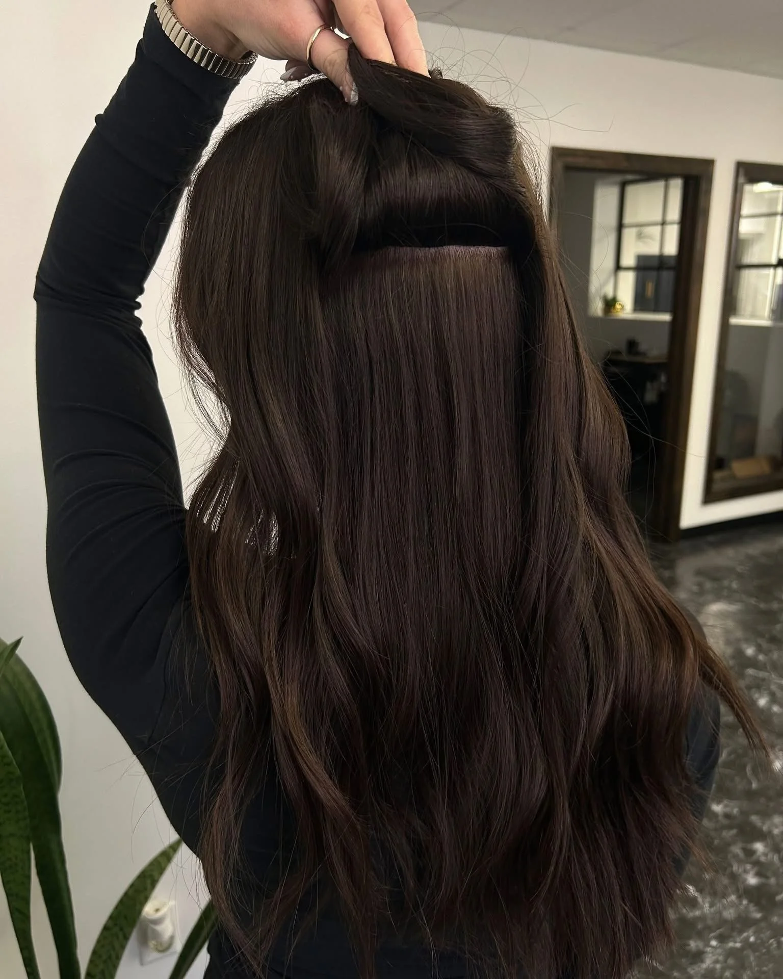 A woman with long, dark brown hair shows a close-up of her hair with a section lifted to reveal a hidden lace frontal hairpiece or wig. She is wearing a black long-sleeve shirt and a silver watch, and it appears she is in a salon or home setting.
