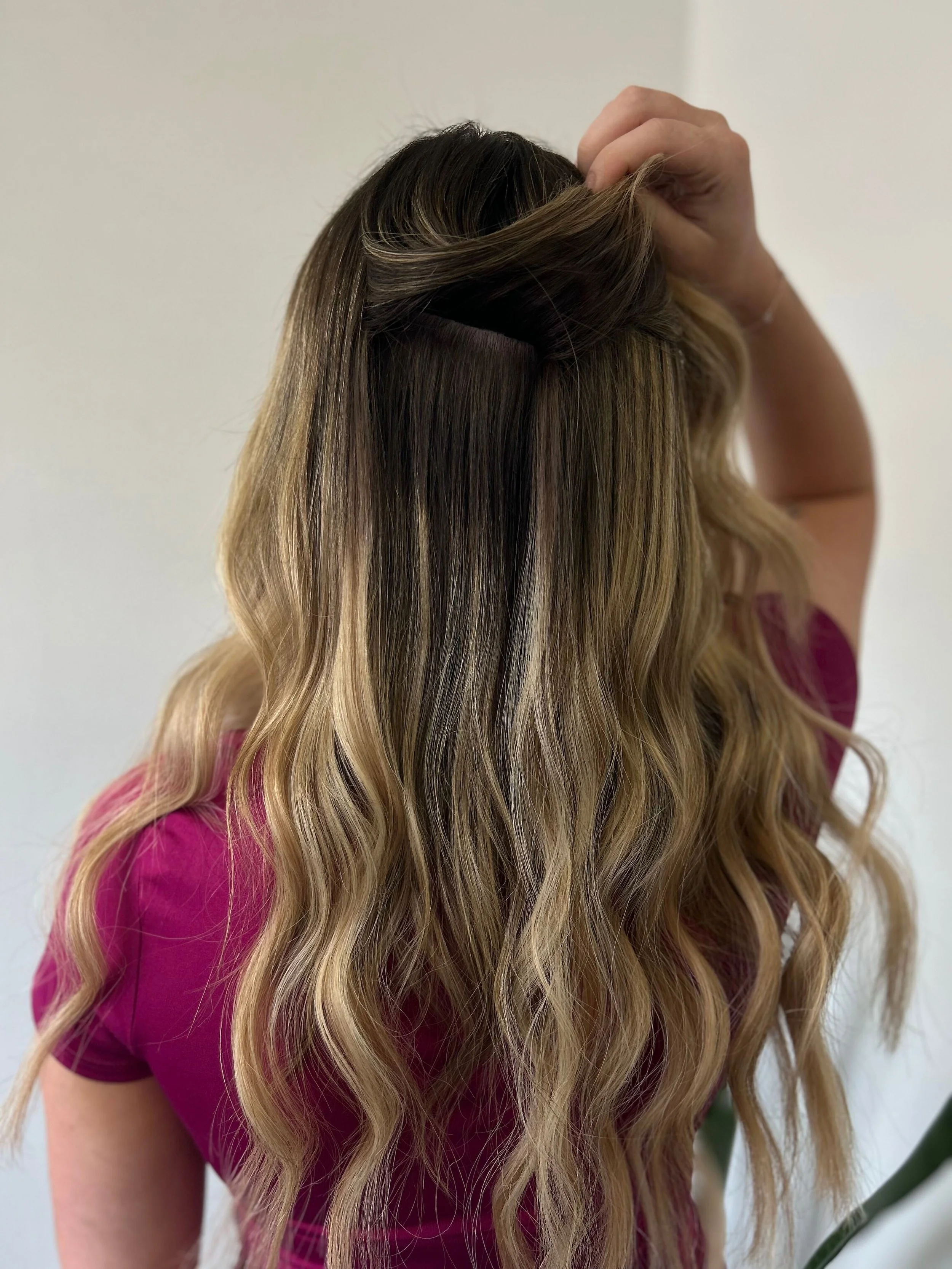 Close-up of a woman with long, wavy, ombre hair in shades of blonde and brown, styling her hair with her hand, wearing a pink top, against a plain white background.