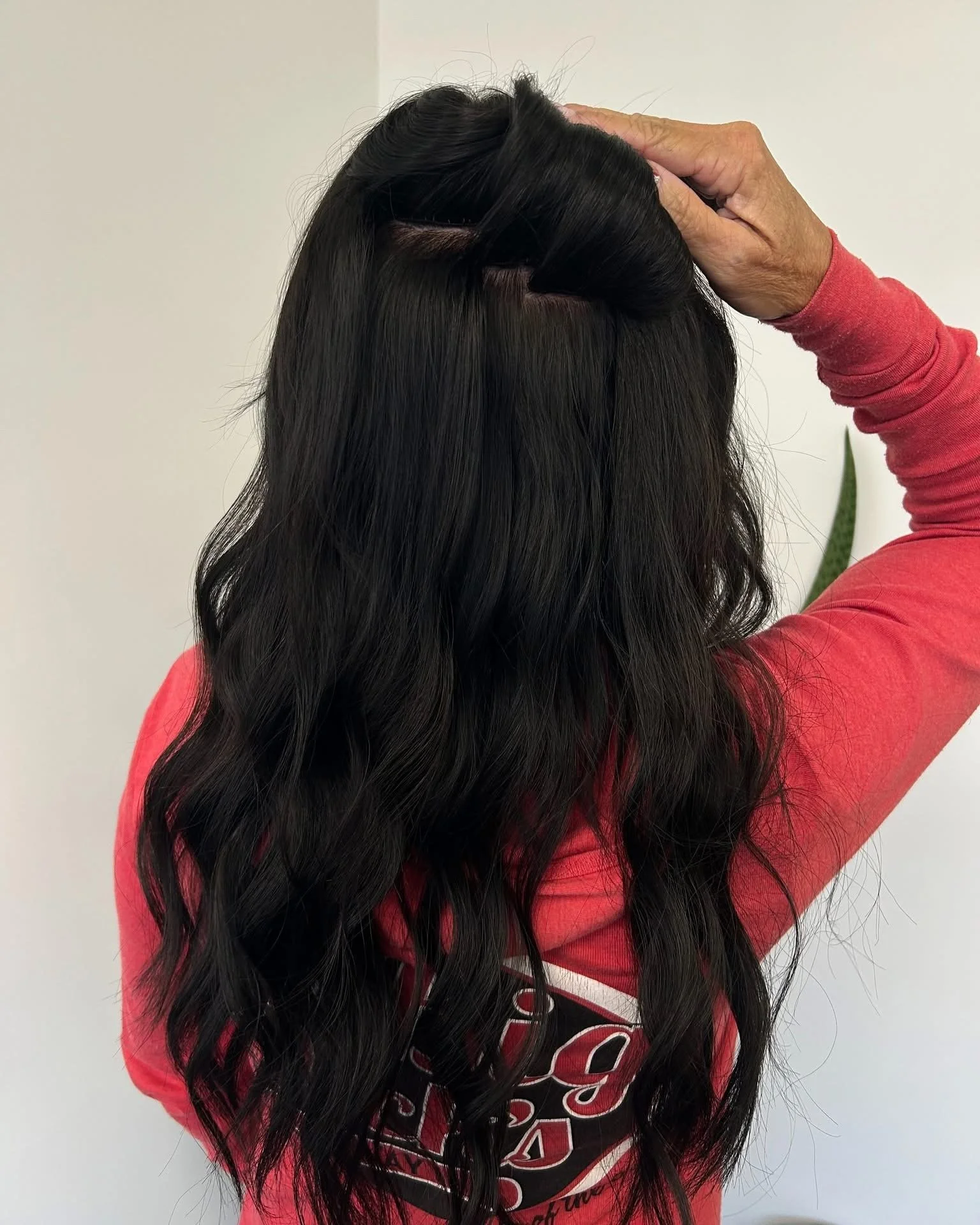Person with long black wavy hair, wearing a red long sleeve shirt, holding their head with one hand.