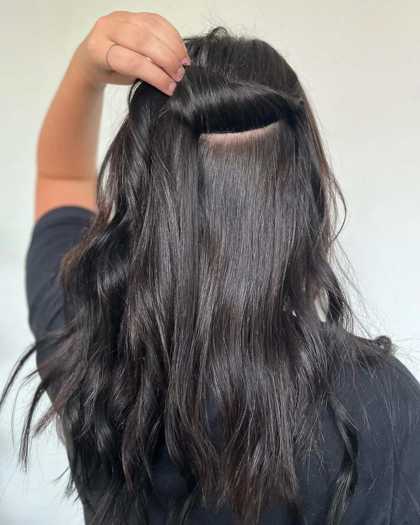 A person with long, dark brown, wavy hair holding up a section to reveal a hair extension or lace wig.