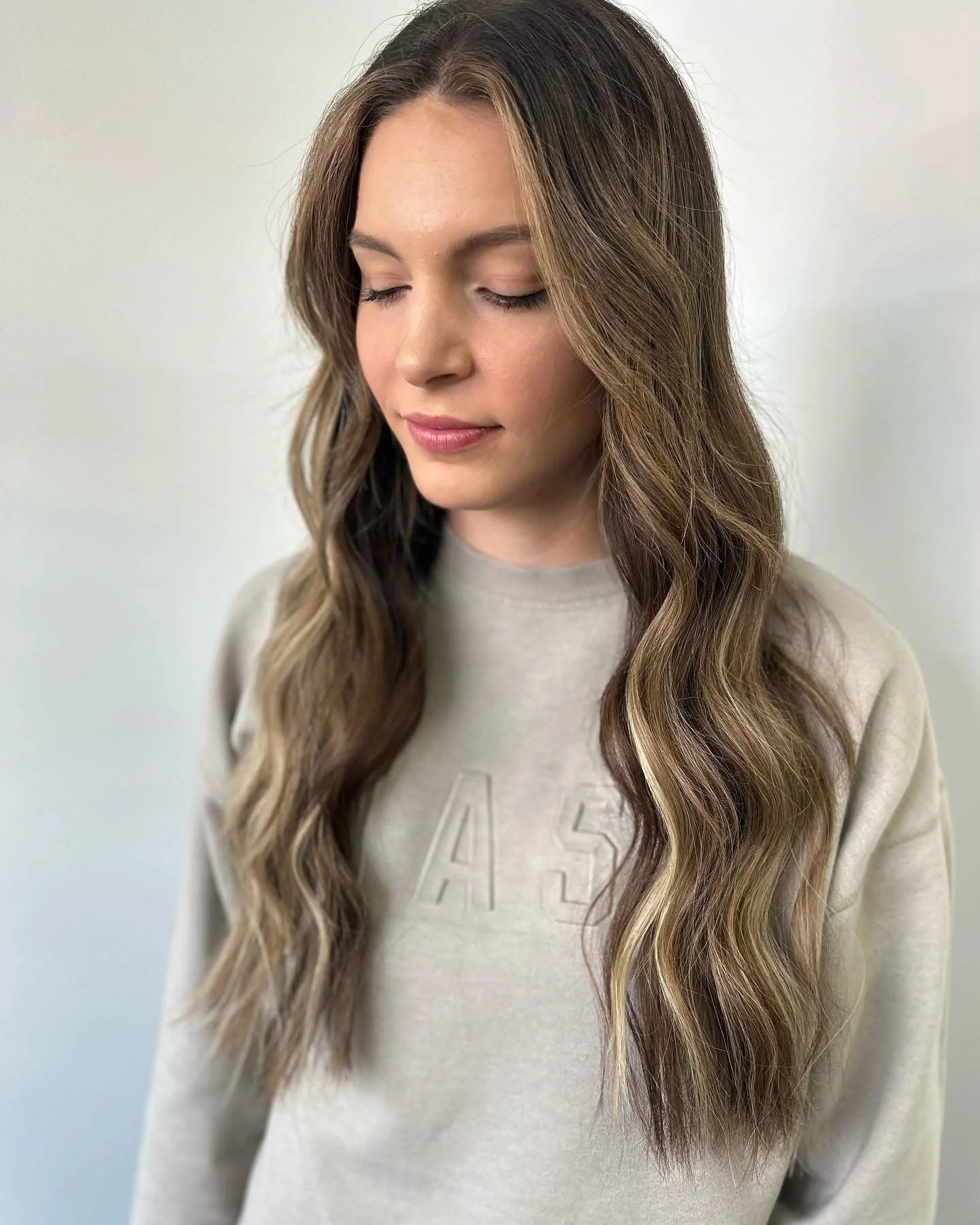 Young woman with long, wavy brown hair with blonde highlights, wearing a beige sweatshirt with embossed letters, and standing against a plain light-colored wall with her eyes closed.