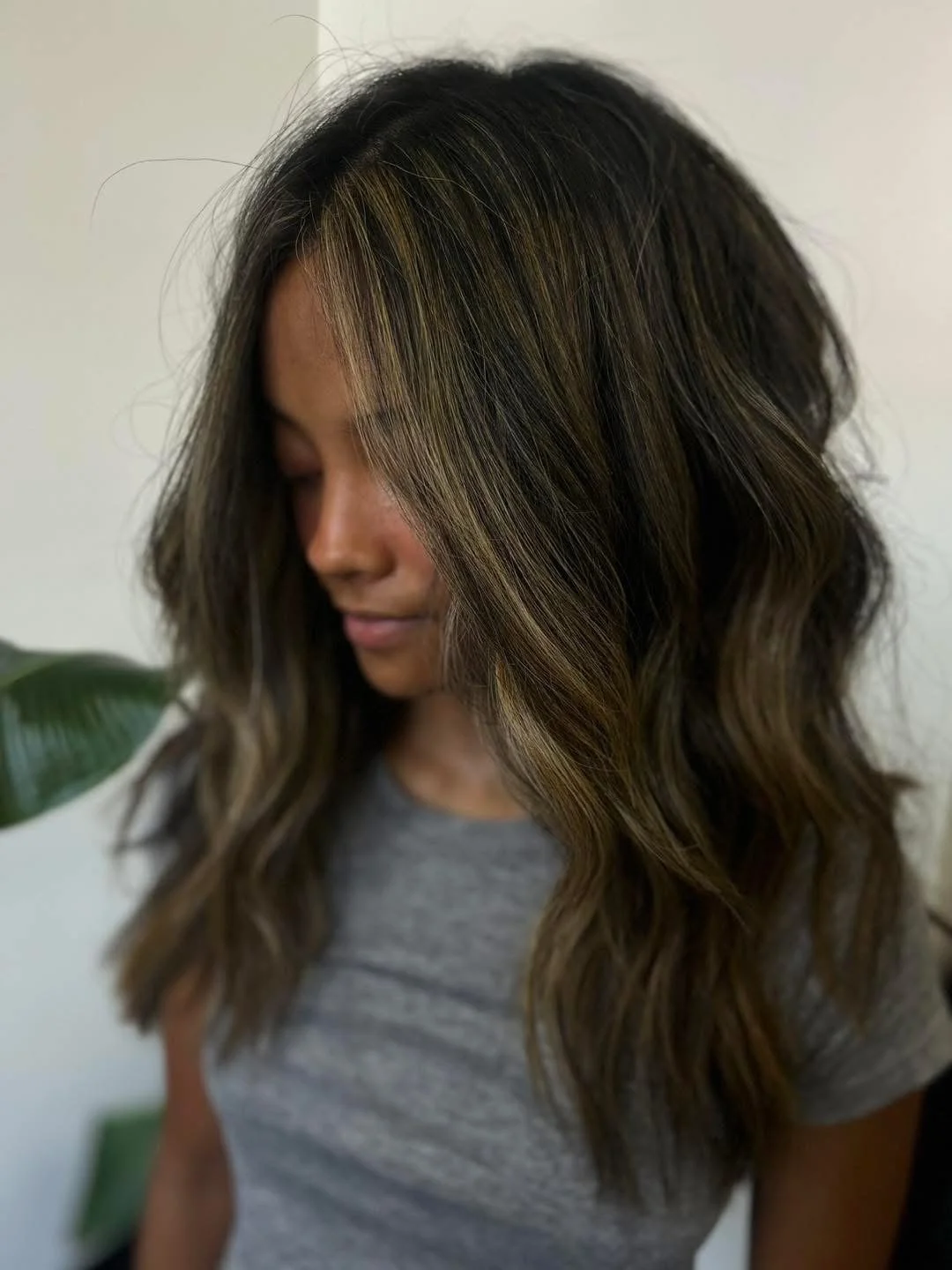 A woman with shoulder-length wavy hair, dark with blonde highlights, wearing a gray t-shirt, looking down with her eyes closed, standing in front of a plain background.