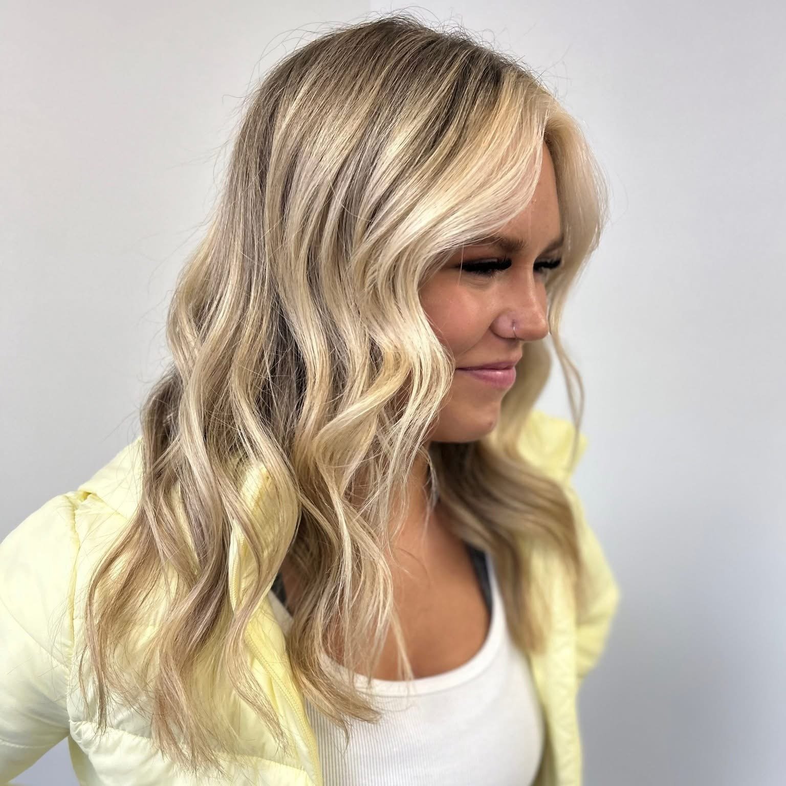 A woman with long wavy blonde hair, wearing a yellow jacket and a white top, standing against a plain white wall, with a slight smile and her eyes closed.