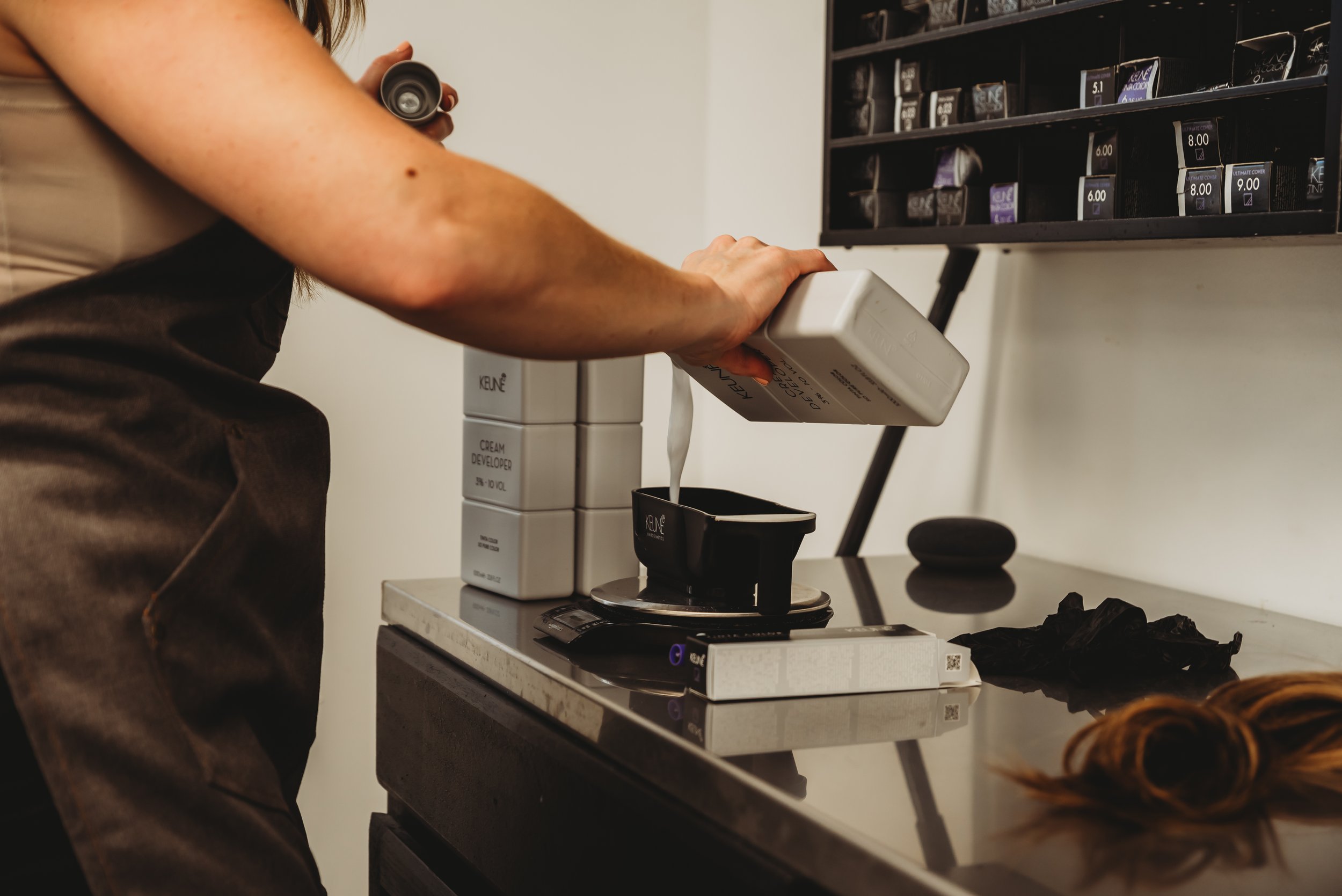 A person pouring liquid from a bottle into a black container on a kitchen counter, with spice jars and a scale nearby.