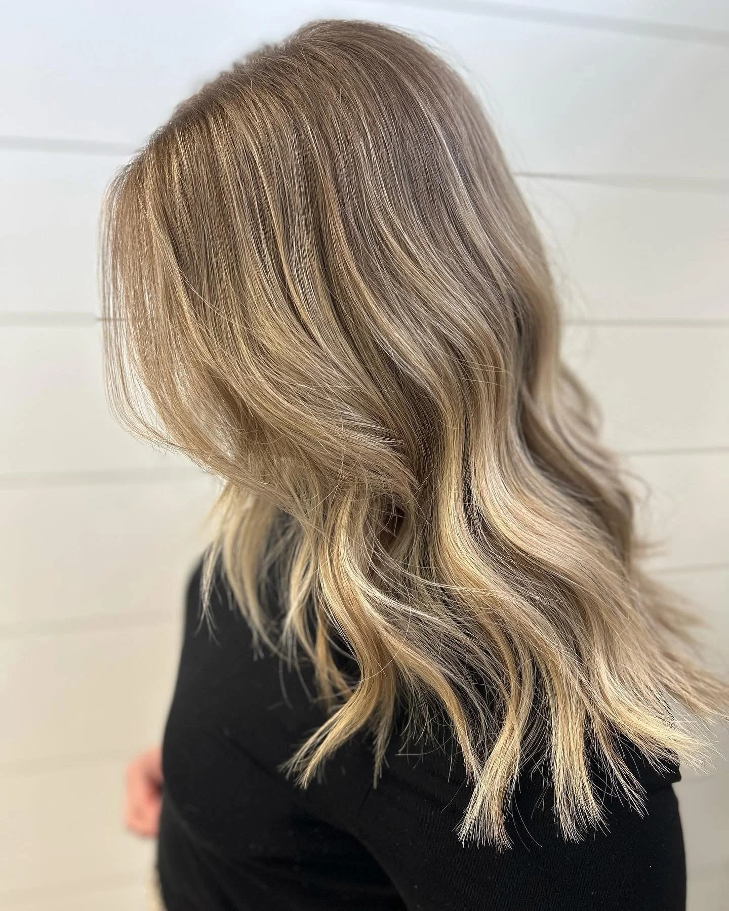 Back view of a woman with long, wavy blonde hair with darker roots, wearing a black top, standing against a white shiplap wall.