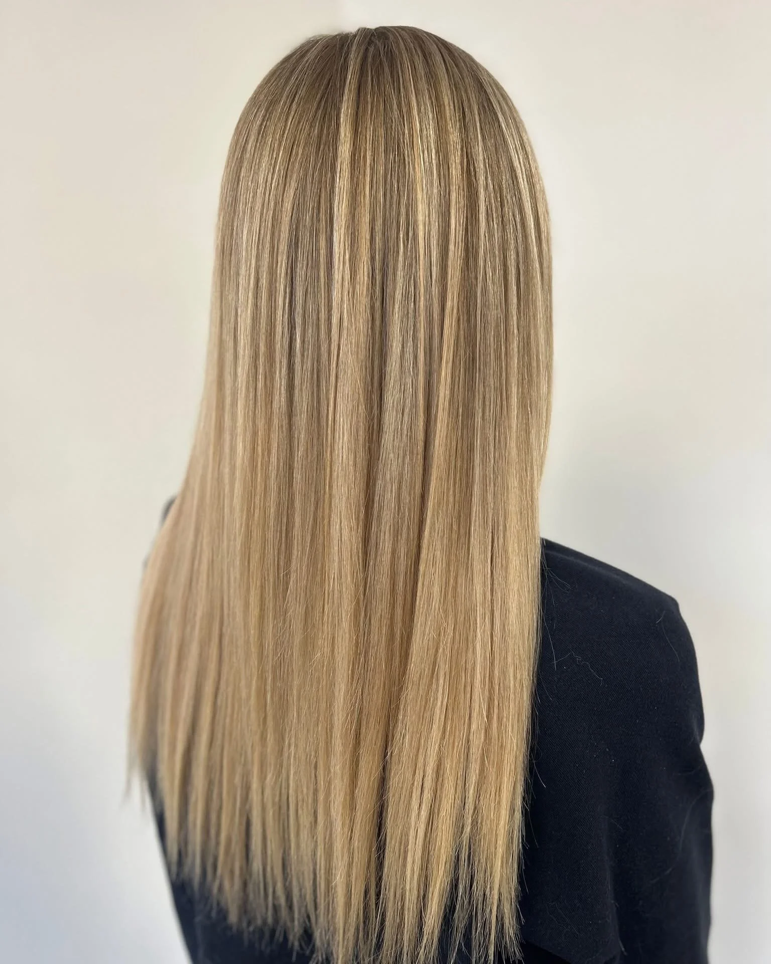 Back view of long, straight, blonde hair with subtle highlights worn by a person dressed in black, standing against a plain light-colored background.