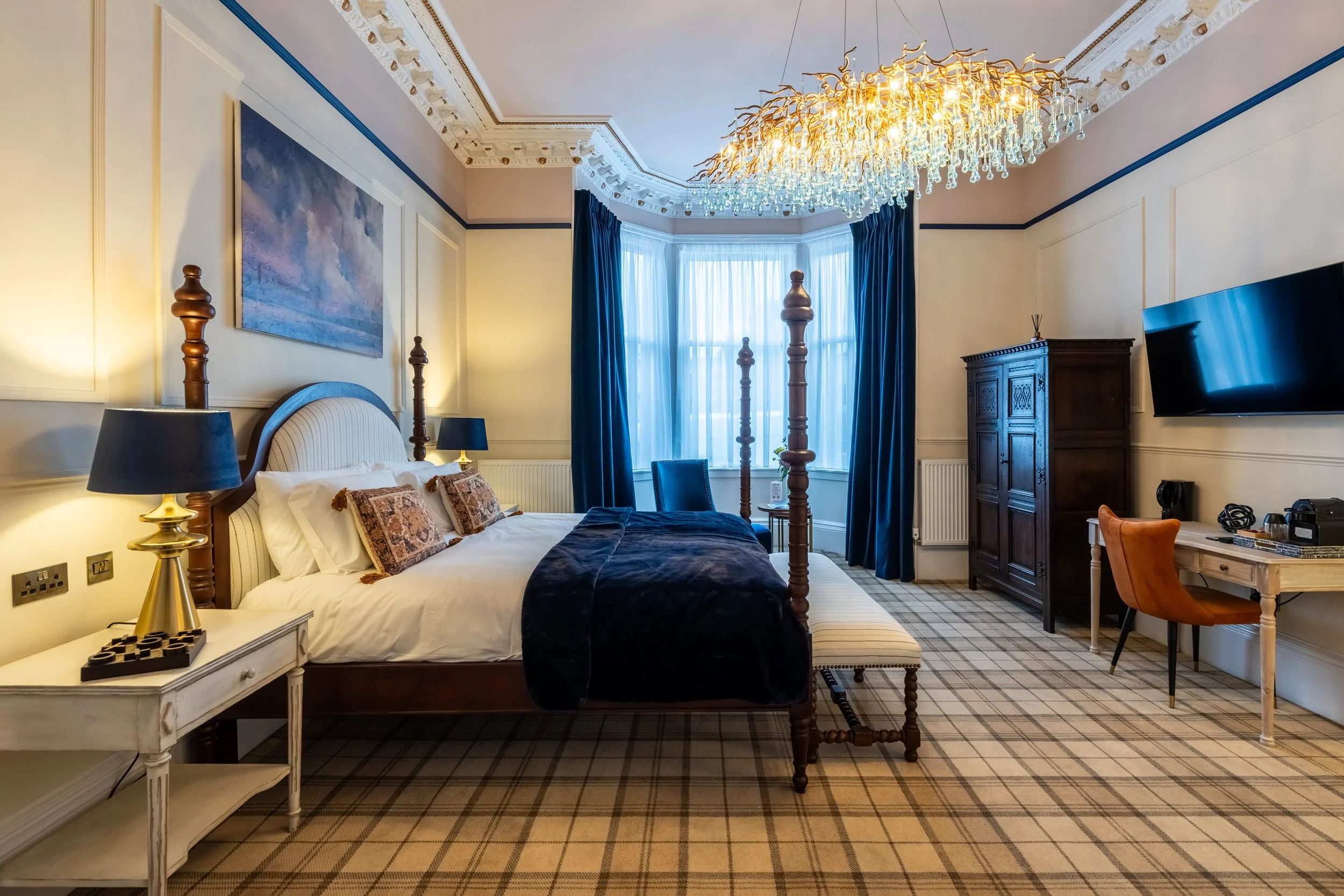 A hotel room with a four-poster bed, blue curtains, a chandelier, a wooden wardrobe, a flat-screen TV, and a desk with a chair.