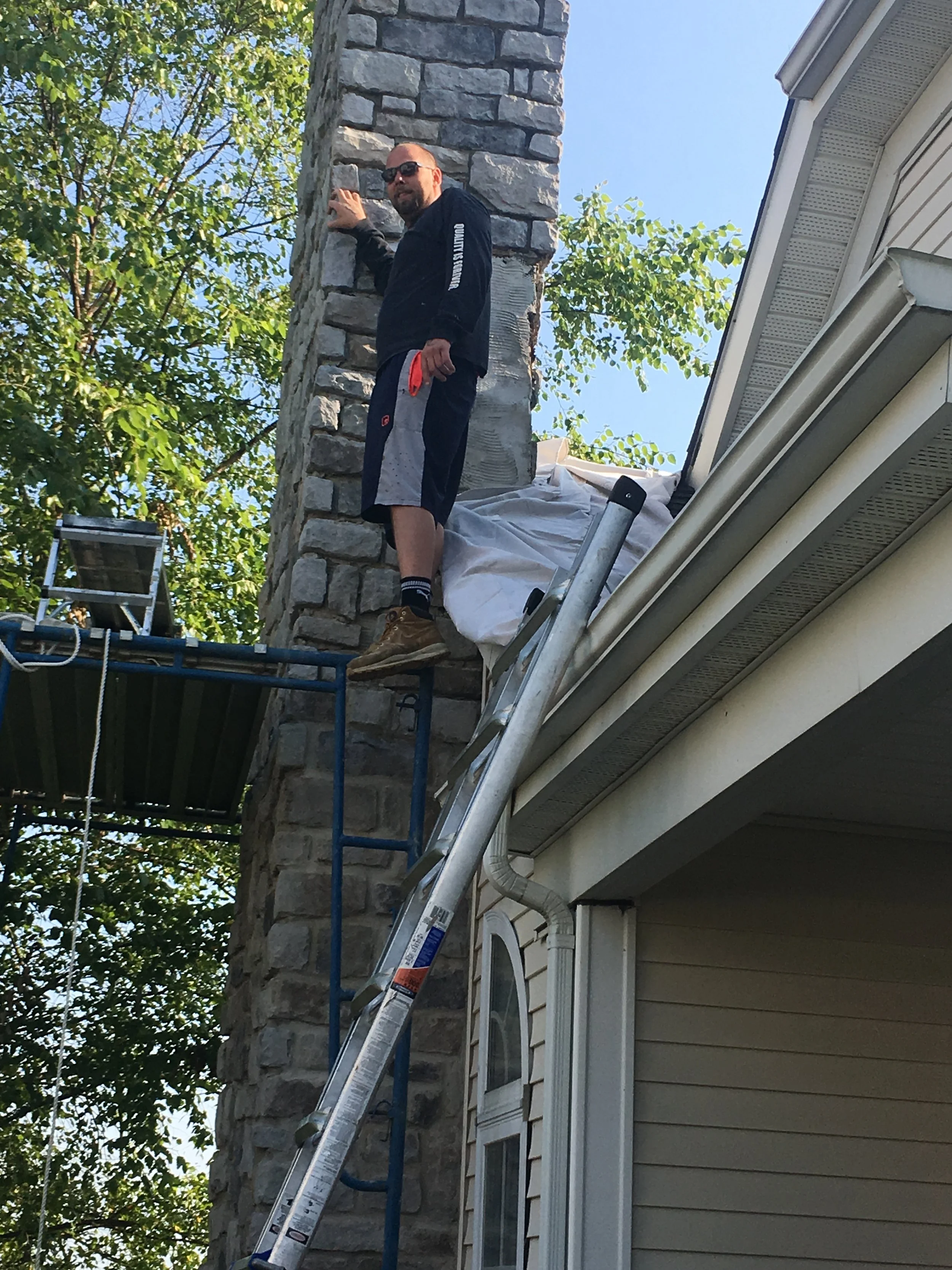 A man standing on a ladder beside a stone chimney, just finished working on building the chimney from stone and cleaning it out and testing it.