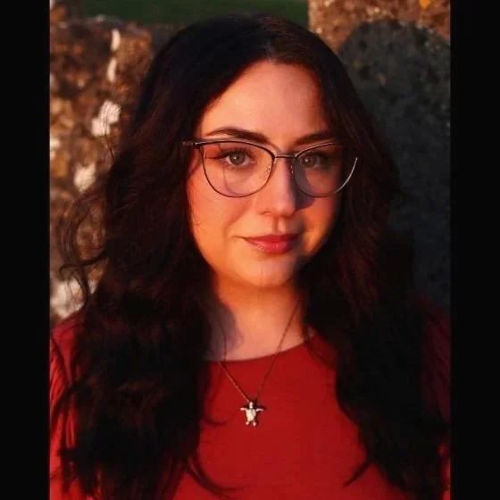 A woman with long dark hair, wearing glasses and a red top, is outdoors during sunset with warm lighting and a rocky background.