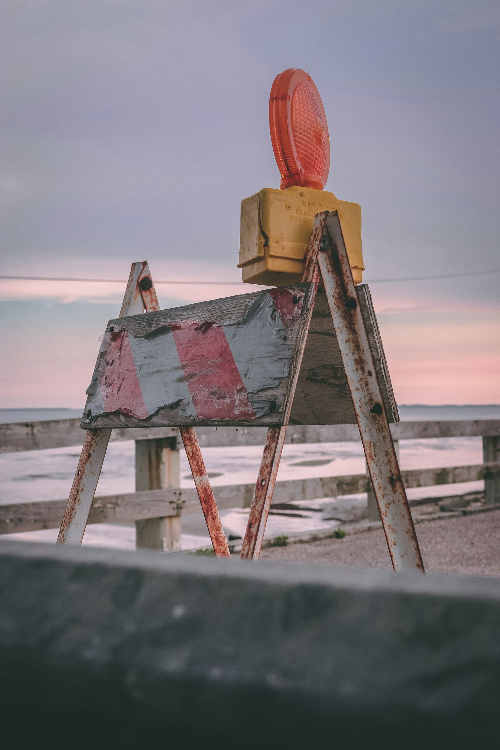 A rusty construction barricade with a worn warning light on top, set by a beach during sunset.