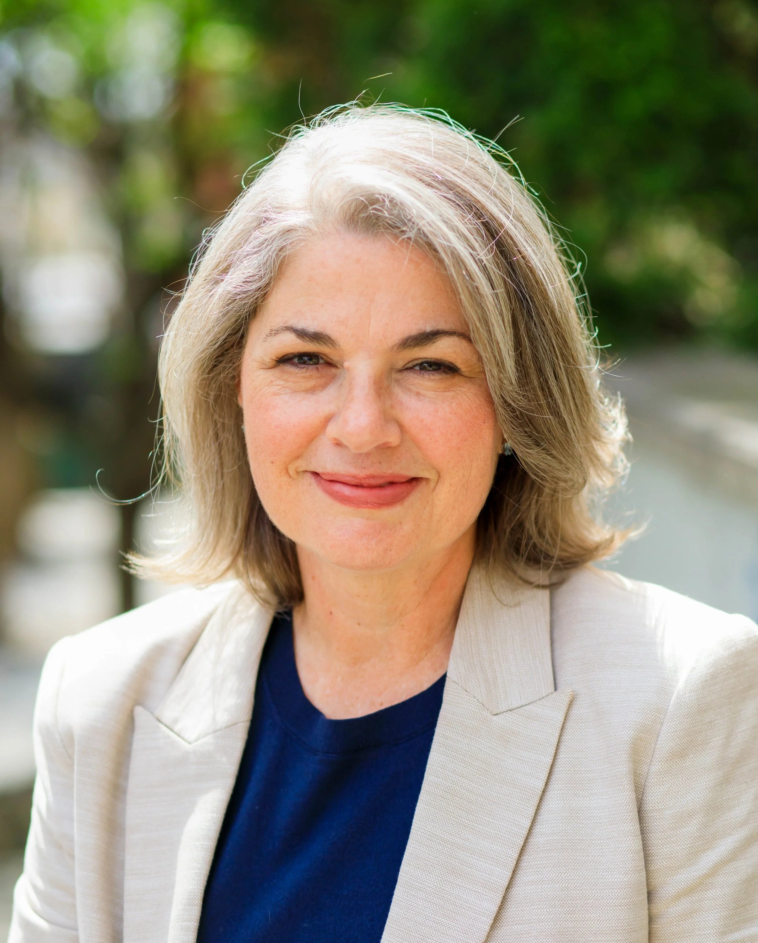 Close-up of a smiling middle-aged woman with gray hair, wearing a beige blazer and navy shirt, outdoors with blurred green foliage in the background.