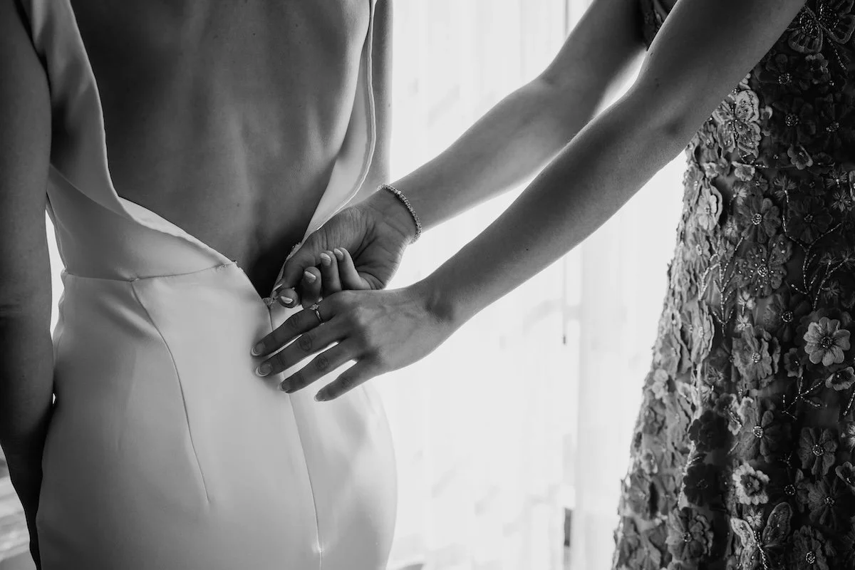 A person in a wedding dress is being helped into the dress by another person, who is zipping up the back of the dress. The scene is black and white.