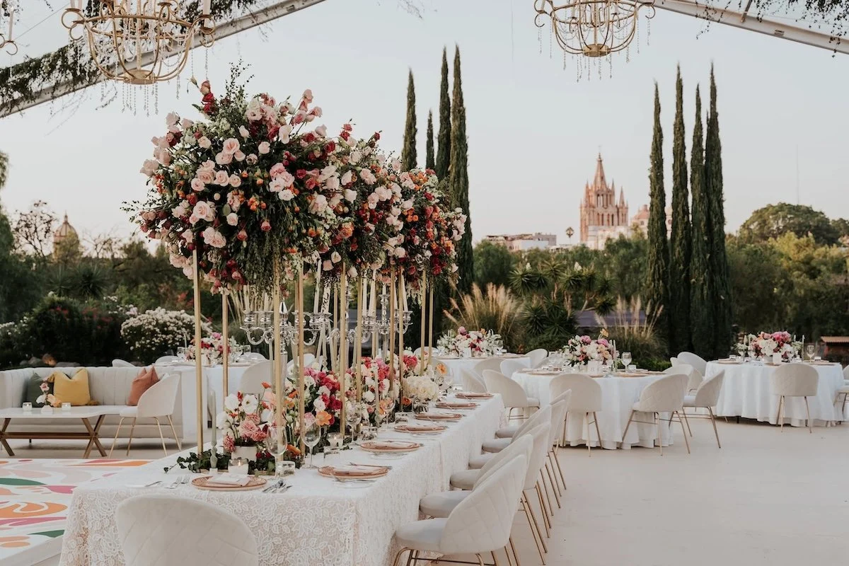 Elegant outdoor wedding reception with round tables and floral centerpieces, set against tall cypress trees and a distant church steeple.