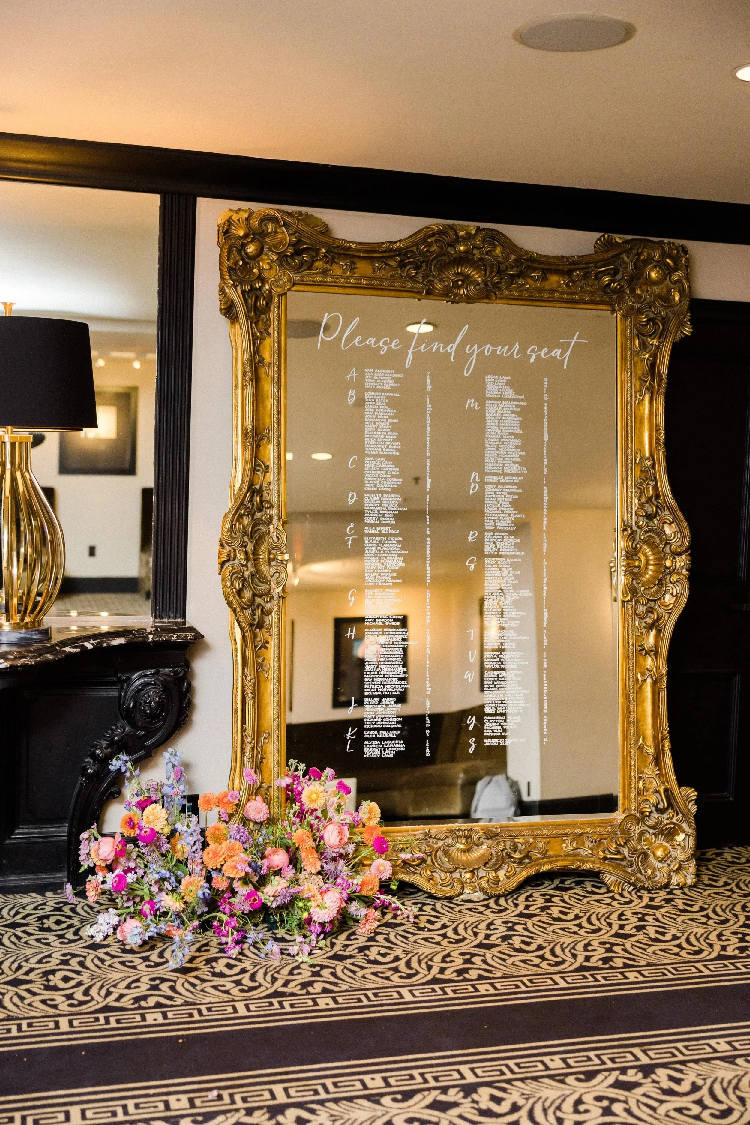 A large ornate gold-framed mirror with a seating chart for an event, possibly a wedding, reflecting a room with lighting. Below the mirror is a bouquet of colorful flowers, including pink, purple, and orange blossoms on a black and gold patterned carpet.