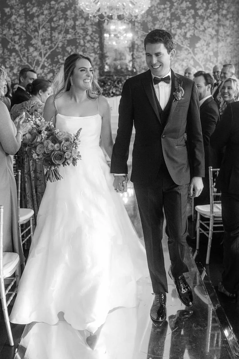 A bride and groom walking hand in hand, smiling at each other during their wedding ceremony surrounded by seated guests.
