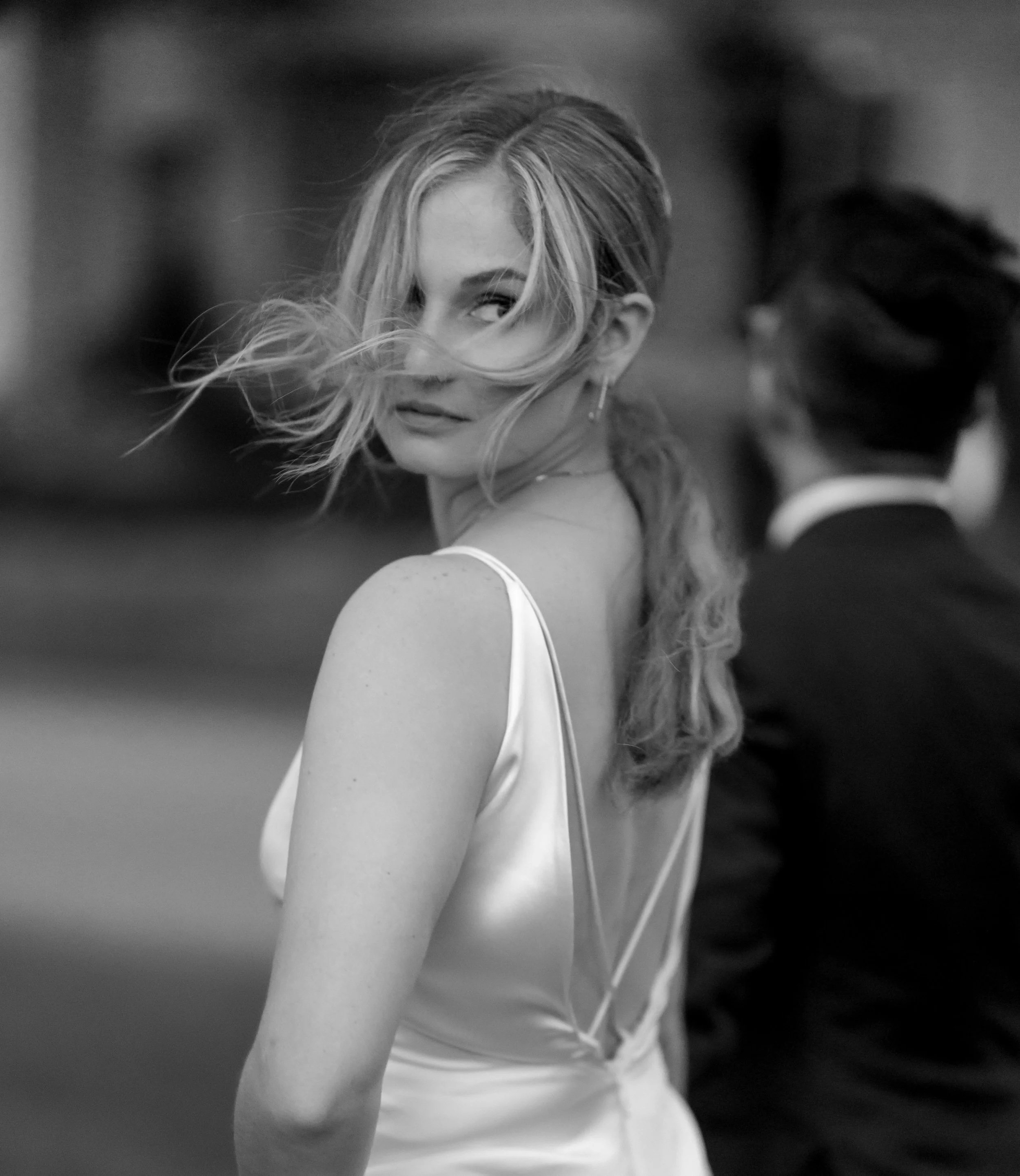 A woman with long hair looks over her shoulder, partially covering her face with her hair, wearing a sleeveless dress, in a black and white photo.