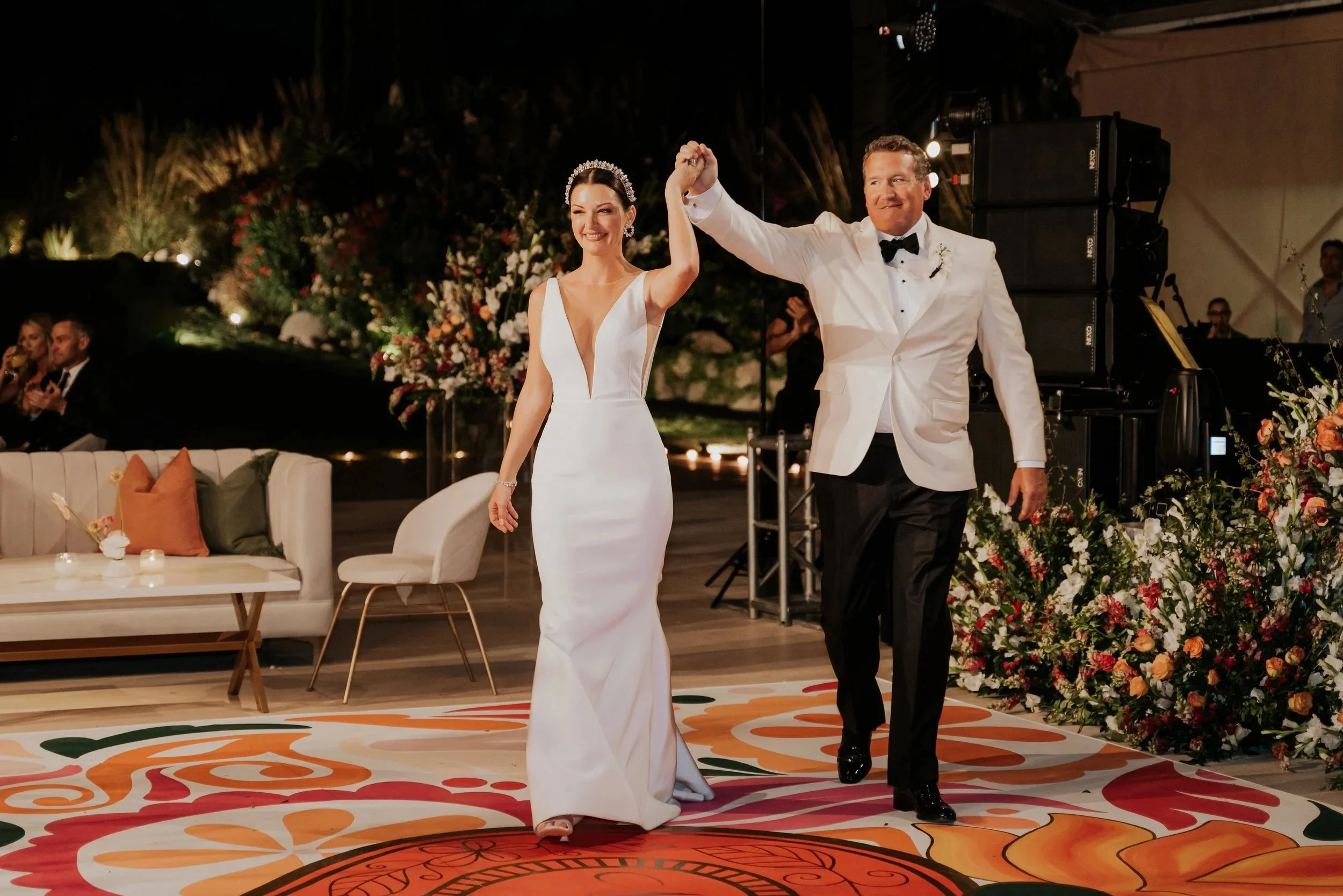 A newlywed bride and groom dancing at their wedding reception. The bride is wearing a white gown with a deep V neckline and a tiara, while the groom is in a white tuxedo jacket with a black bow tie. They are smiling and holding hands. The background features floral arrangements, a band or DJ setup, and guests watching.