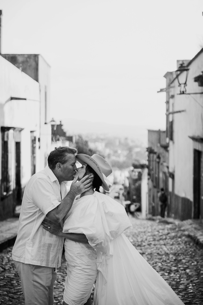 A black and white photo of a couple sharing a kiss on a cobblestone street wearing light-colored clothing, with the woman in a large-brimmed hat, in a small historic town.