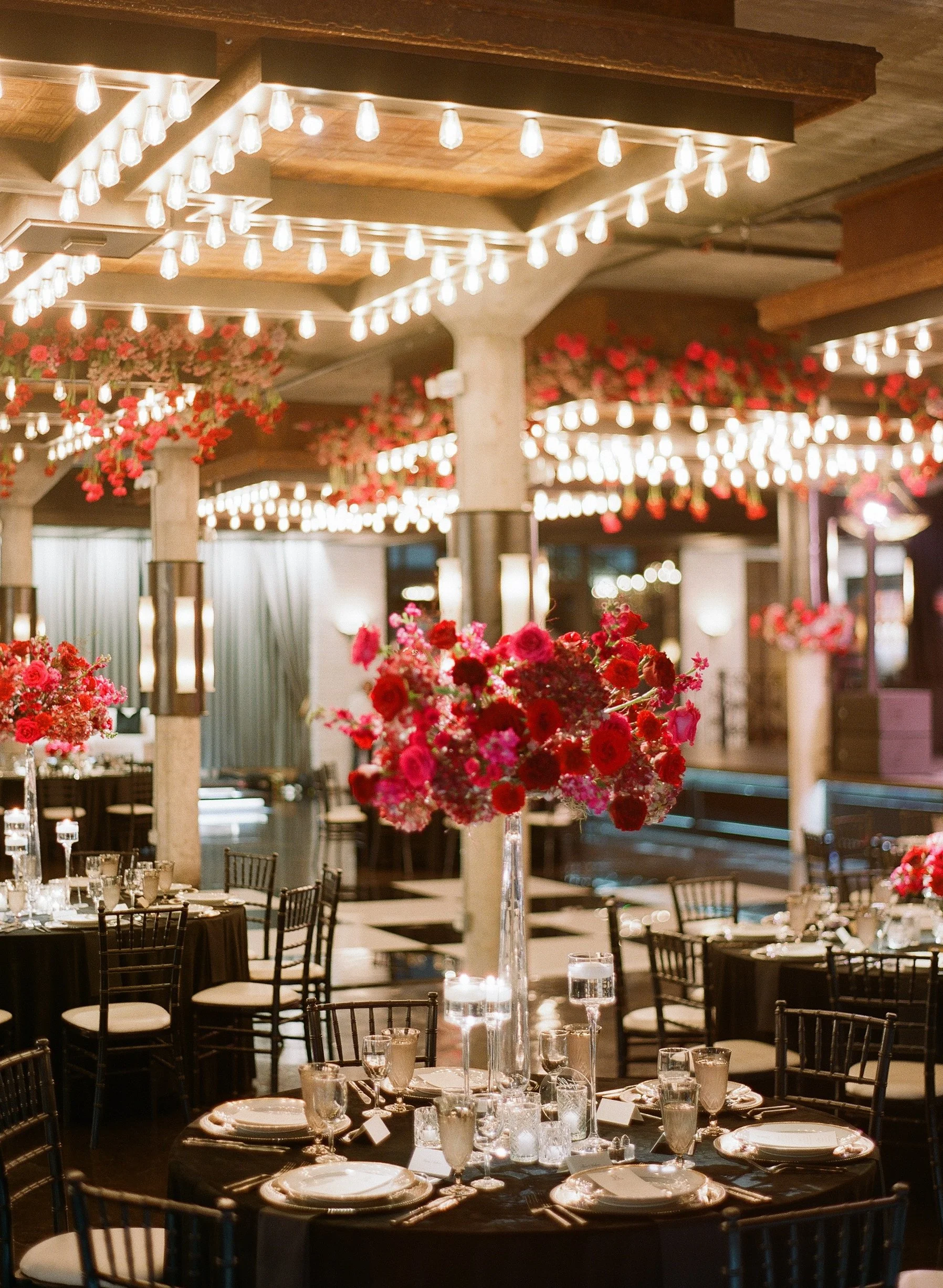 Elegant banquet hall decorated for an event with round tables, black tablecloths, floral centerpieces, and string lights hanging from the ceiling.
