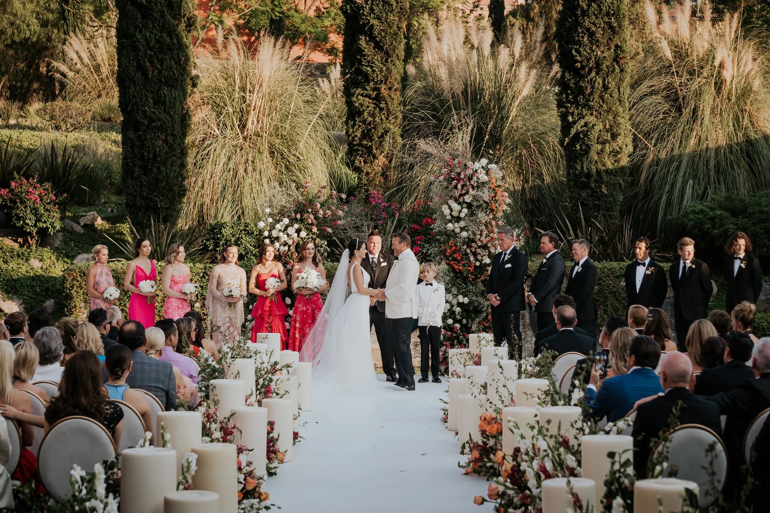 A wedding ceremony taking place outdoors with a couple at the center exchanging vows. Guests seated on both sides, surrounded by lush greenery and tall plants, with floral arrangements decorating the background.